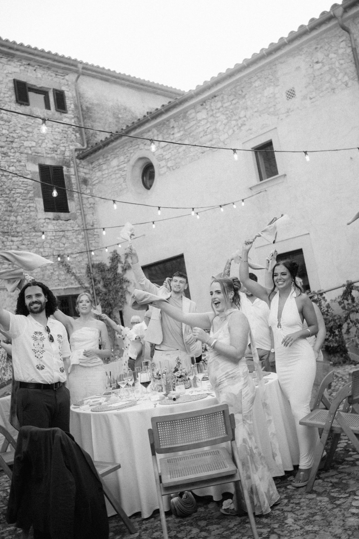 Black and white photo of wedding guests celebrating joyfully at outdoor reception tables.