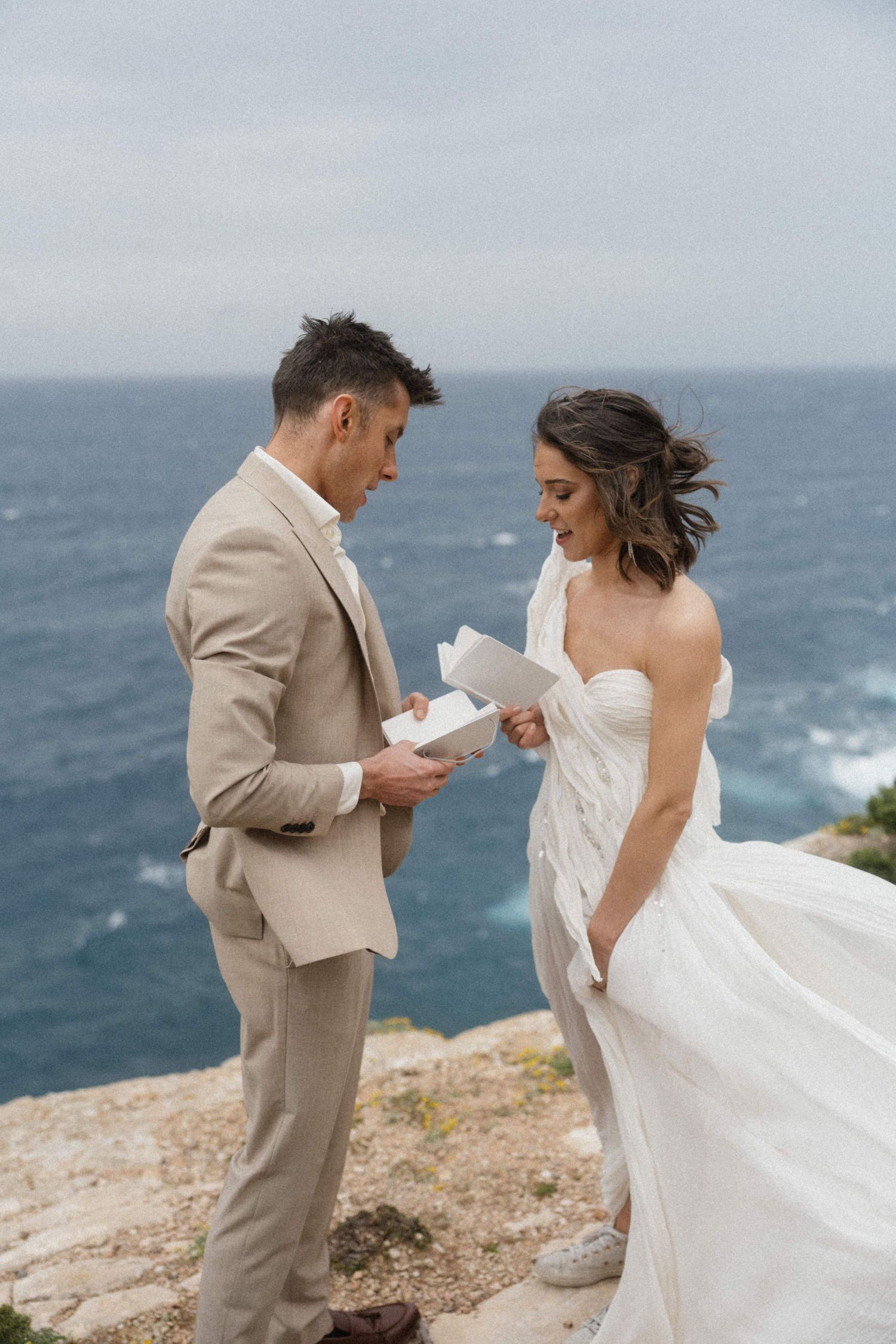 Bride and groom exchange vows on a rocky cliffside overlooking the ocean, she in a white gown, he in a beige suit.