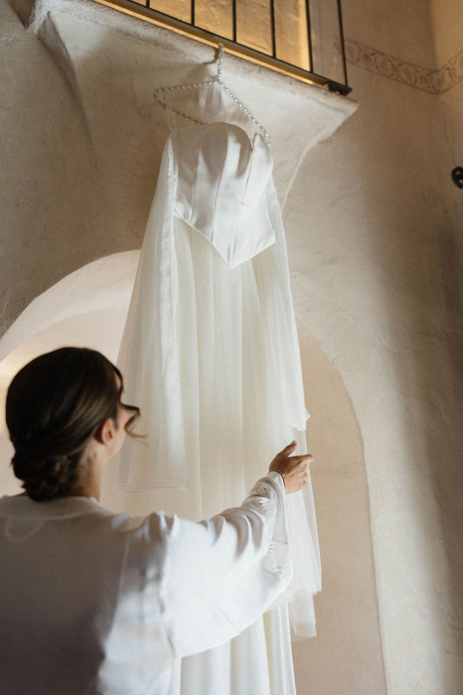 Bride in white robe reaching for elegant wedding dress hanging on arch in rustic venue.