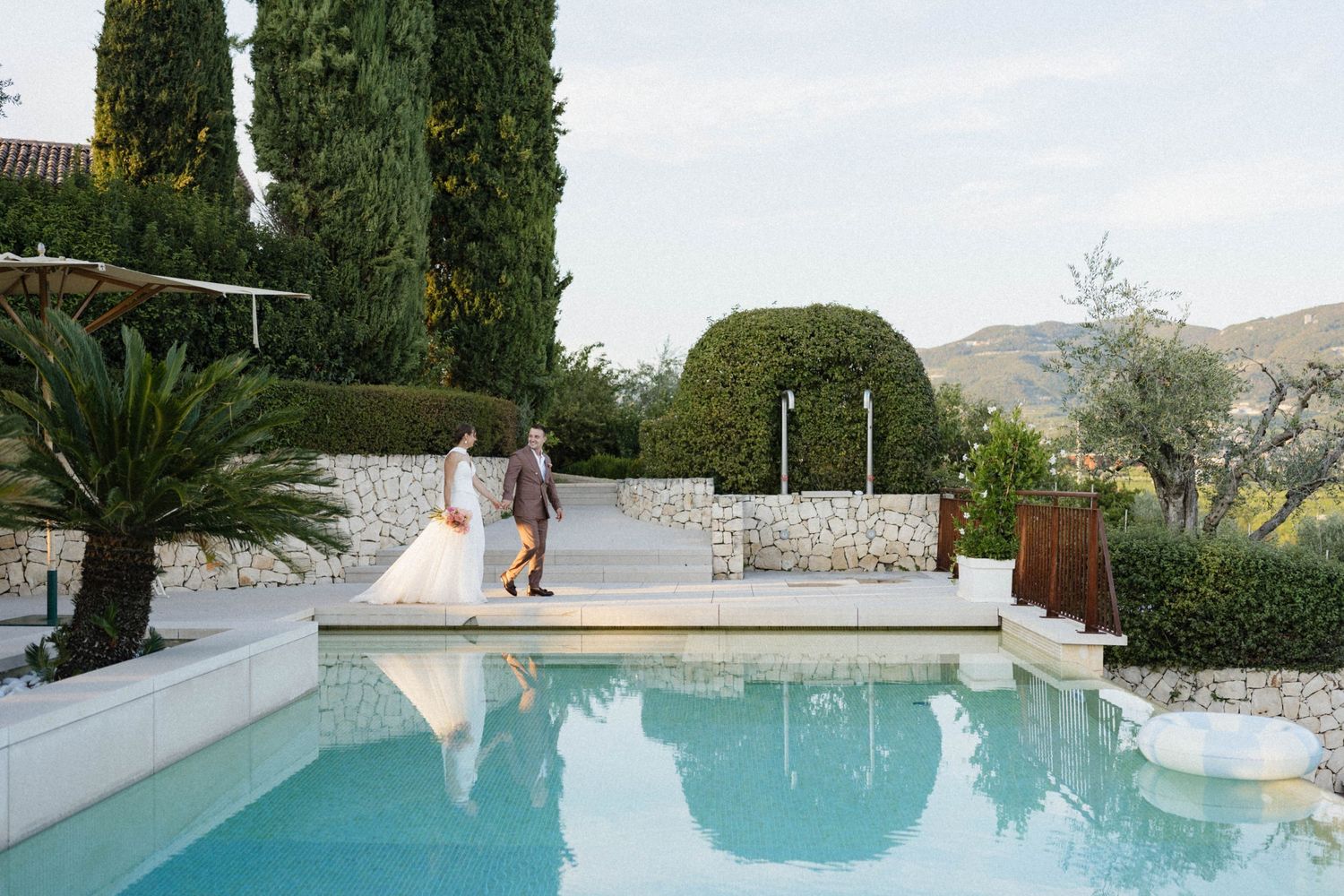 Bride and groom walking beside a luxury pool with cypress trees and manicured hedges at an elegant wedding venue.