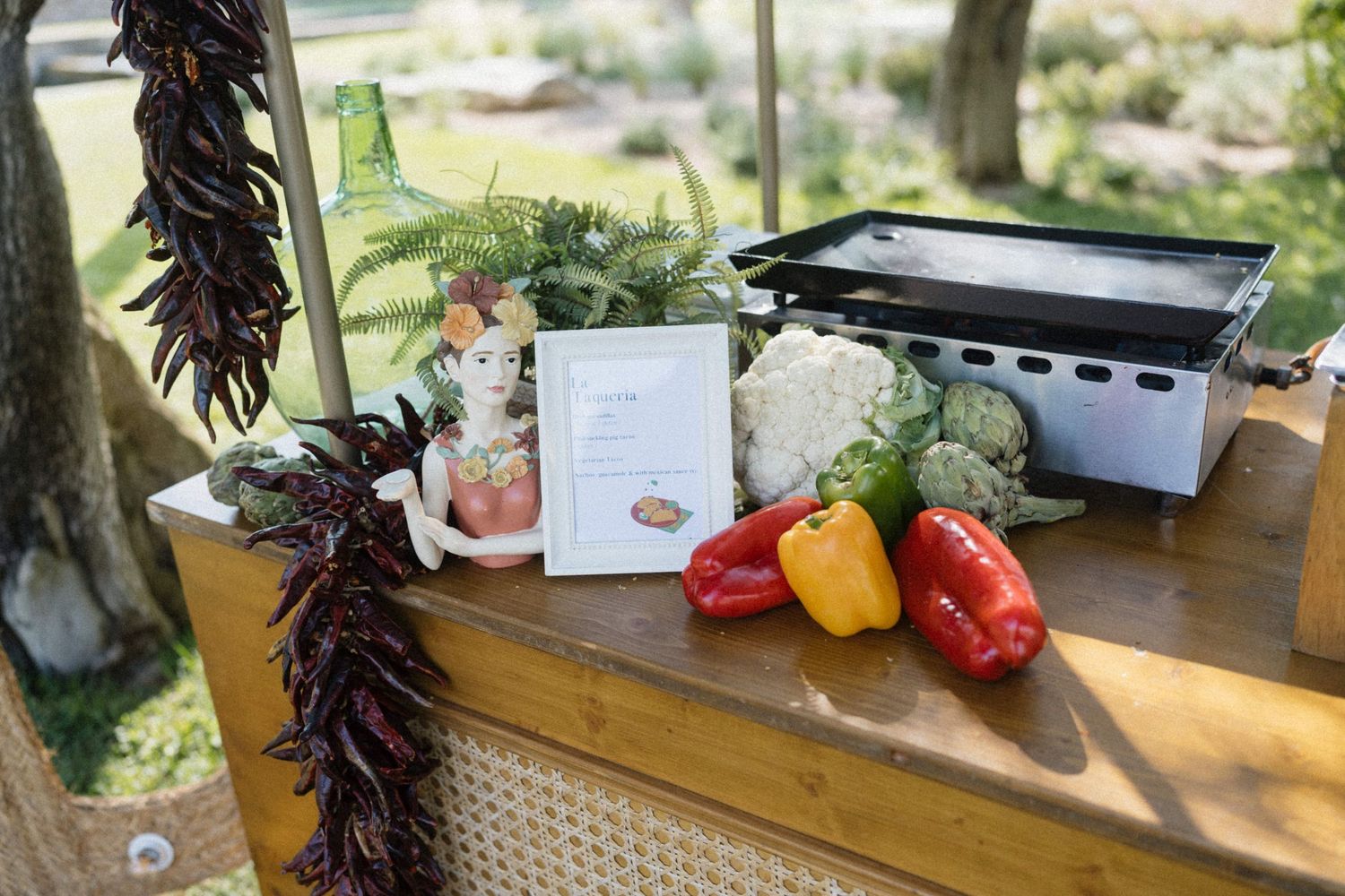 Outdoor cooking station with colorful bell peppers, cauliflower, dried chili pepper garland, and a portable grill on a wooden table.