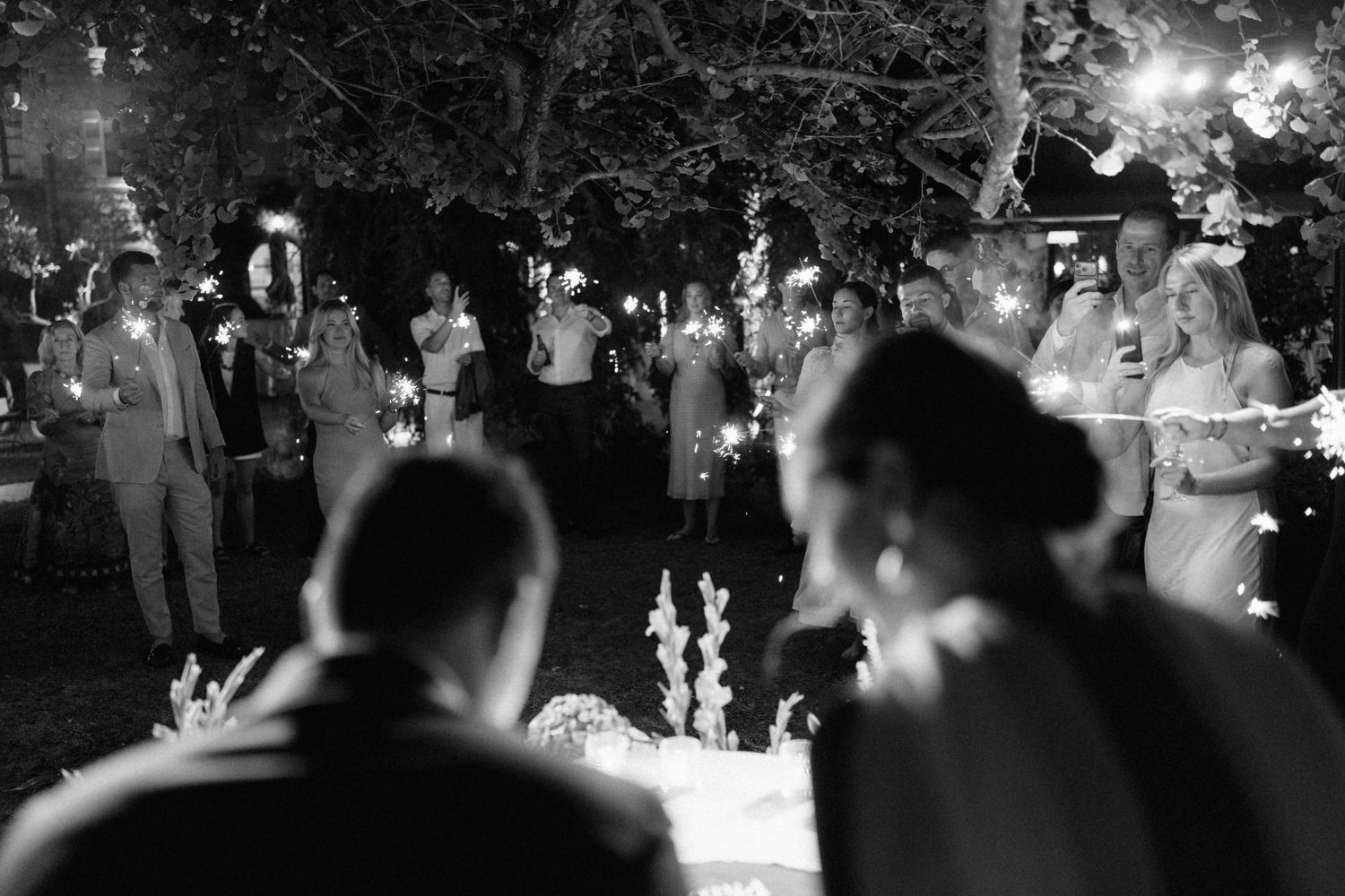 Black and white wedding reception photo with guests holding sparklers outdoors at night, bride and groom seated in foreground.