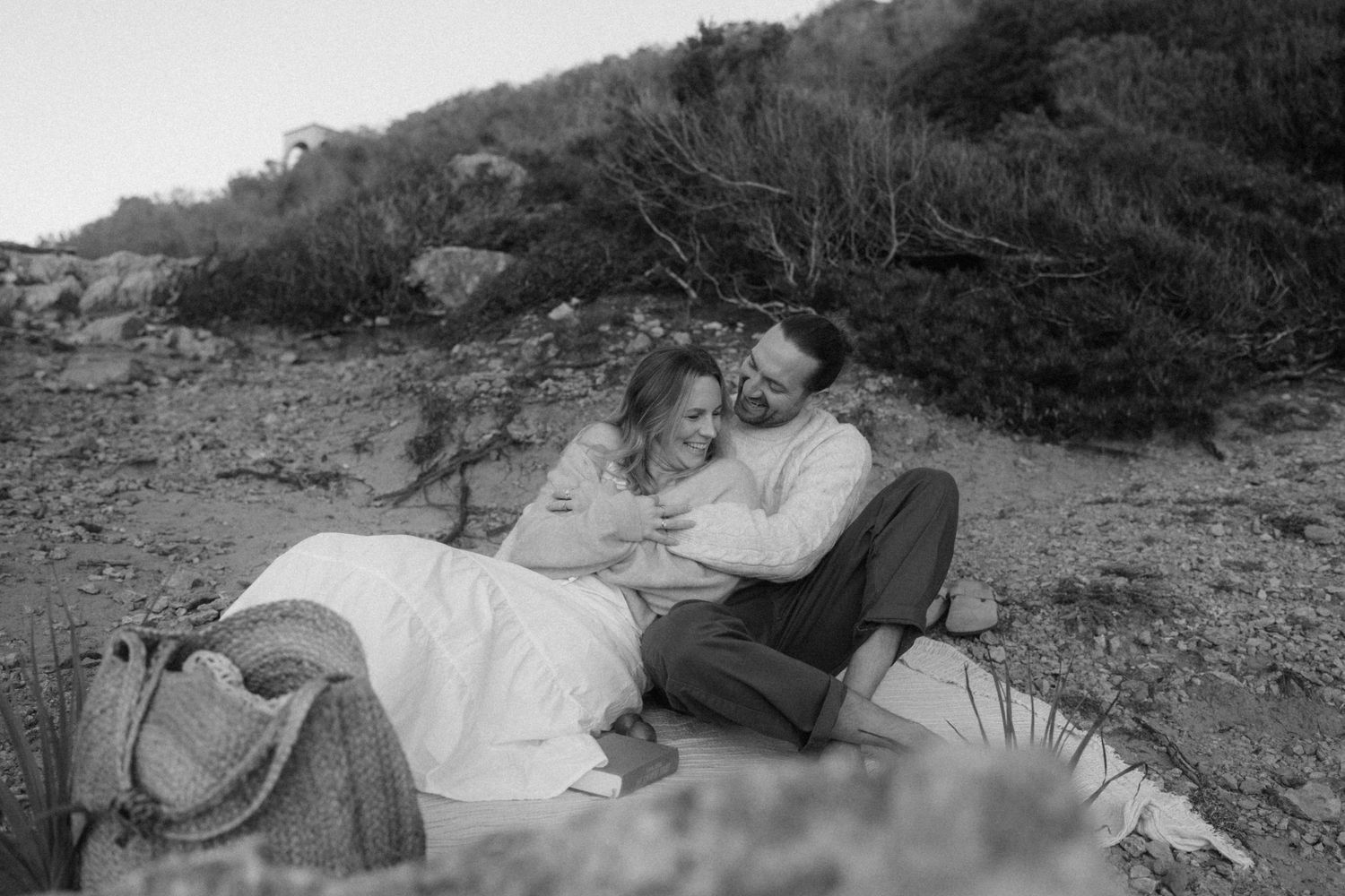 Black and white photo of a couple in wedding attire cuddling on a sandy beach surrounded by natural greenery.