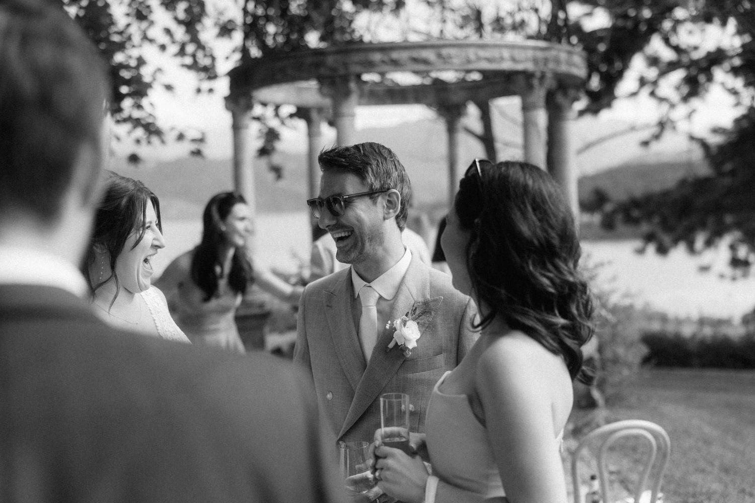 Black and white photo of groom laughing with guests during outdoor wedding cocktail hour.