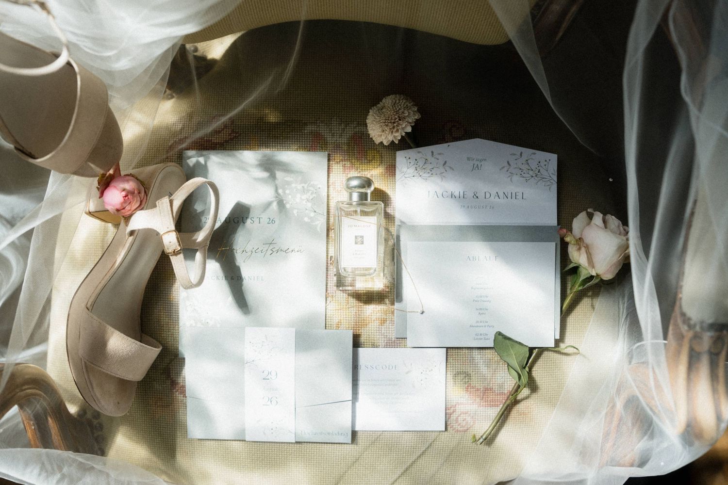 Elegant wedding flat lay with Jo Malone perfume, floral invitations, nude heels, roses, and sheer veil in soft natural light.