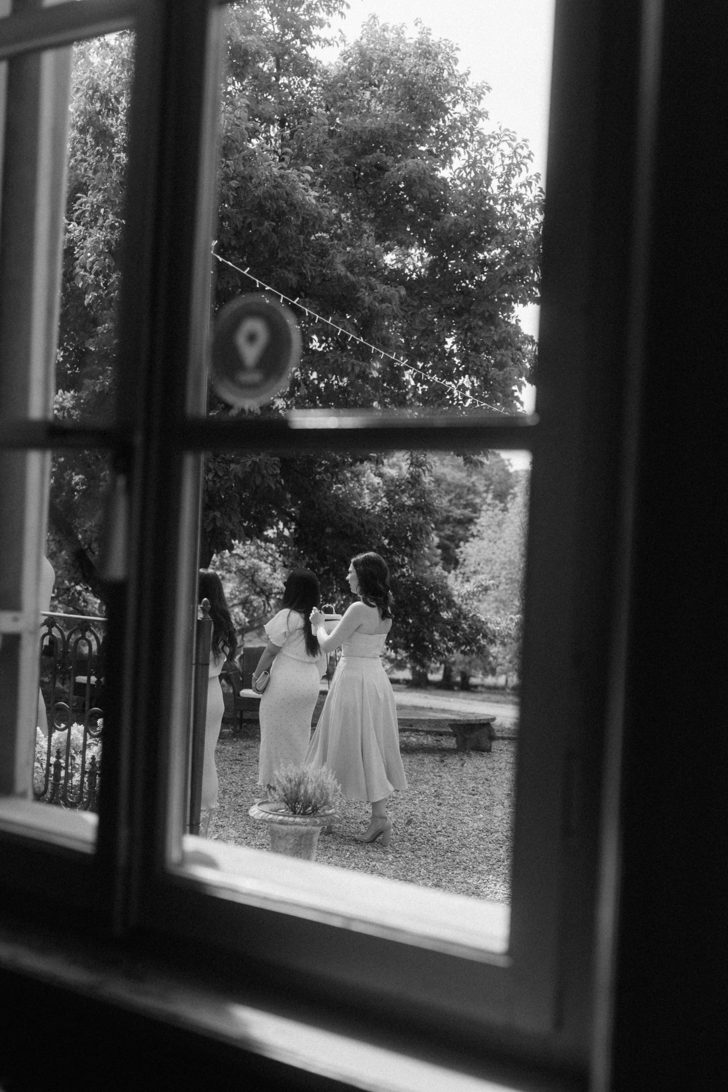 Two bridesmaids in white dresses viewed through a vintage window at an outdoor wedding venue.