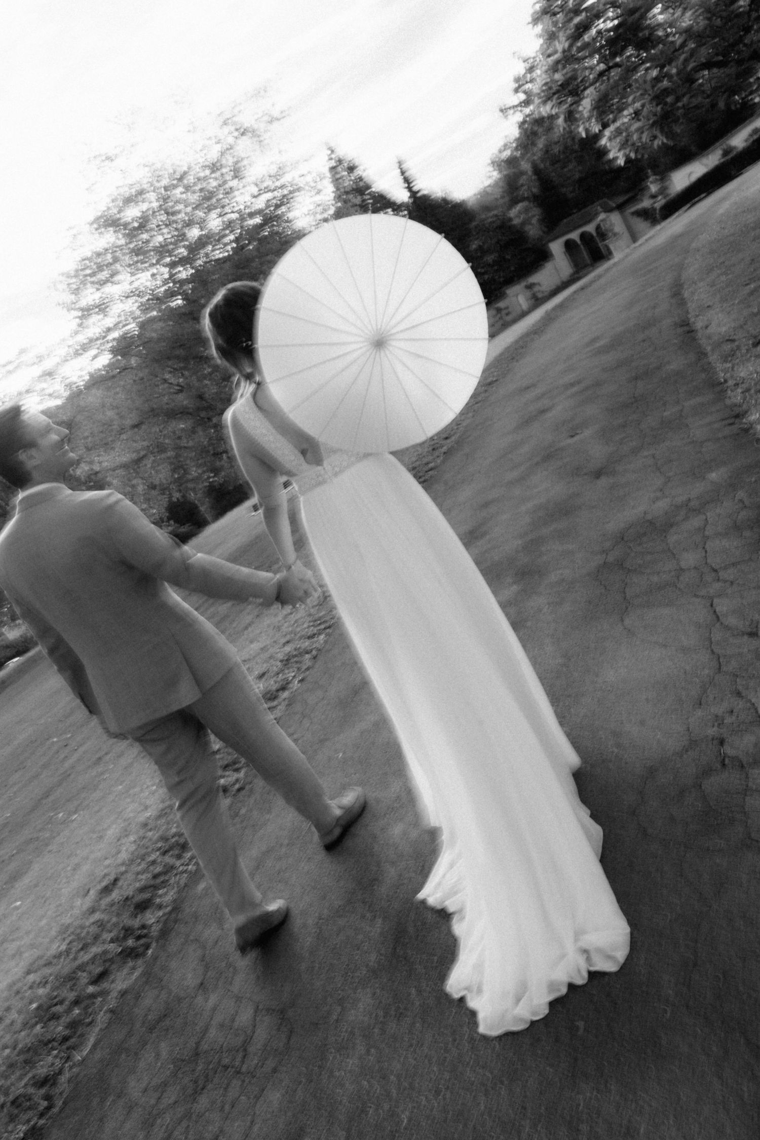 Black and white photo of bride and groom walking with white parasol umbrella at outdoor wedding.