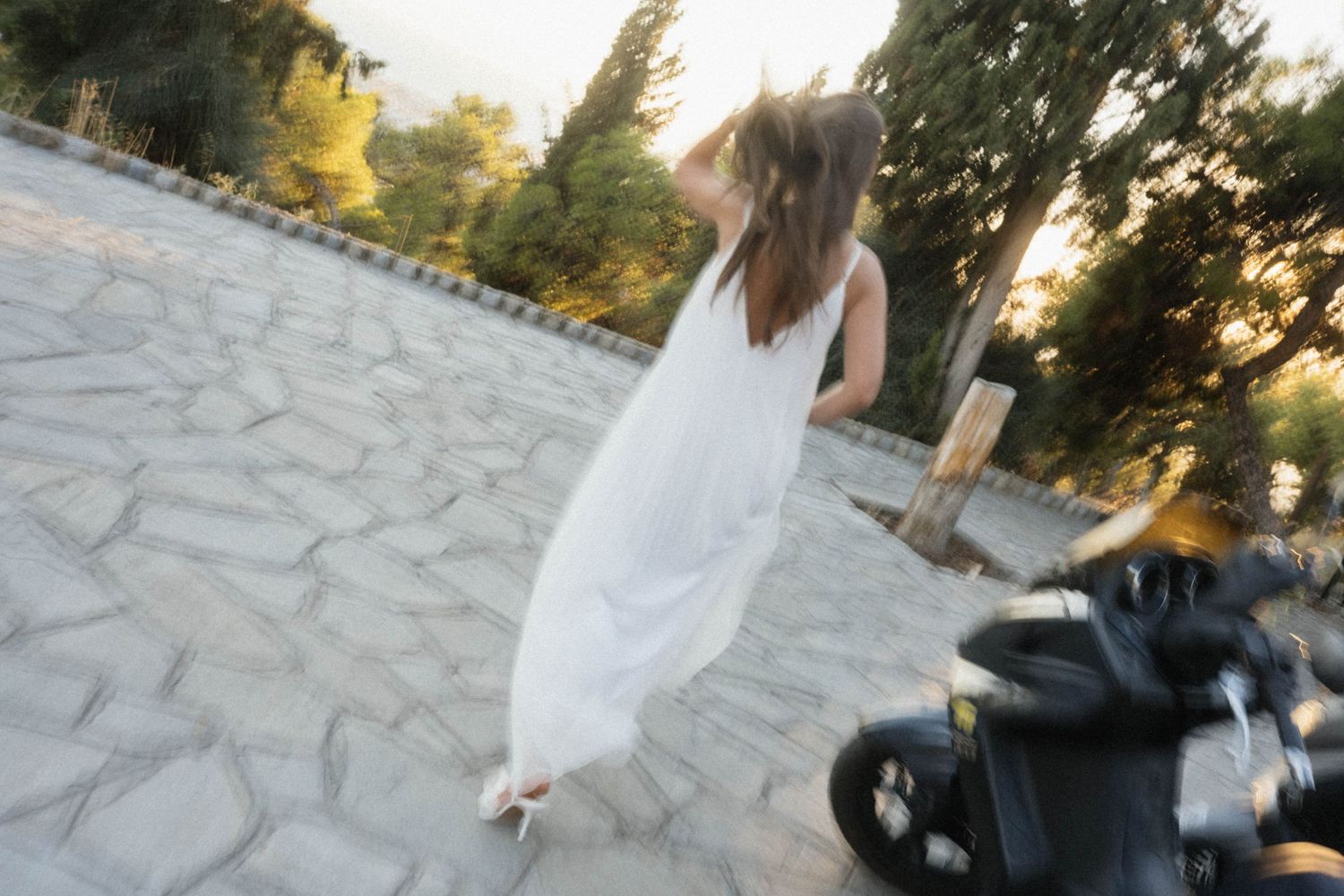Woman in white dress walking near a motorcycle on a stone paved path surrounded by trees at sunset.