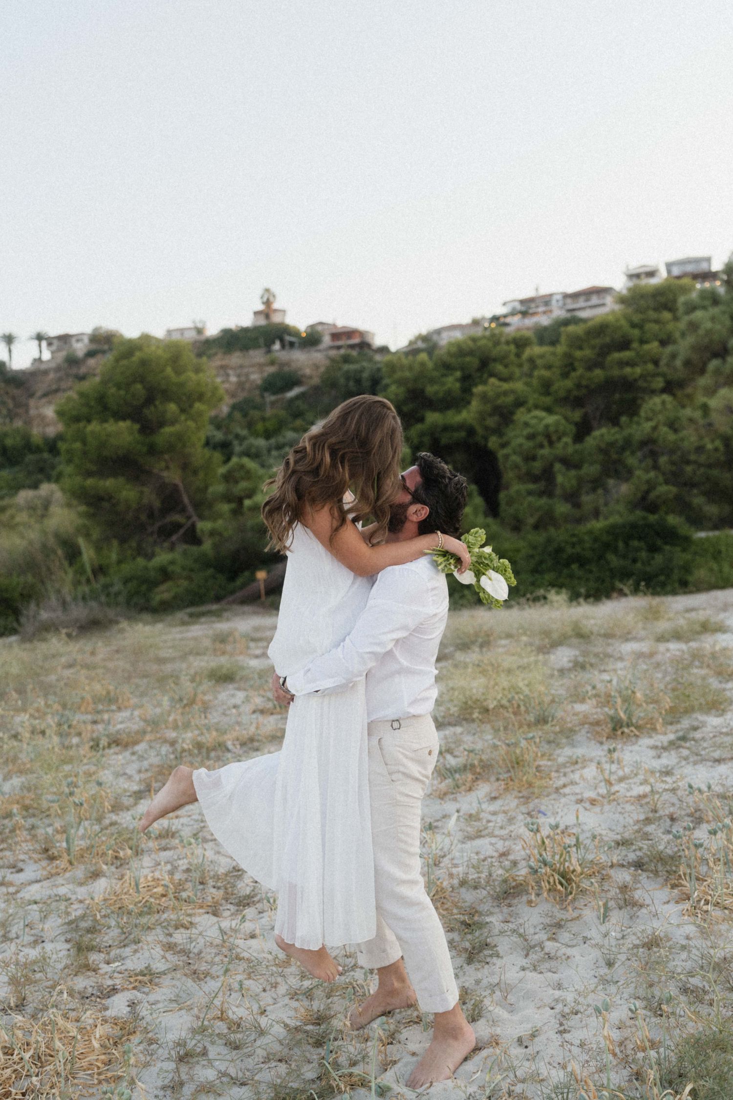 Couple in white embracing and kissing outdoors on sandy ground with lush green trees and hillside homes in background.