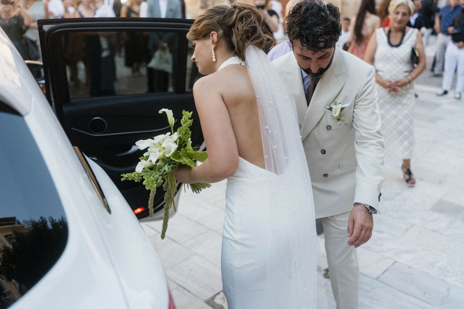 Bride in white gown holding bouquet with groom in beige suit entering car after wedding ceremony.