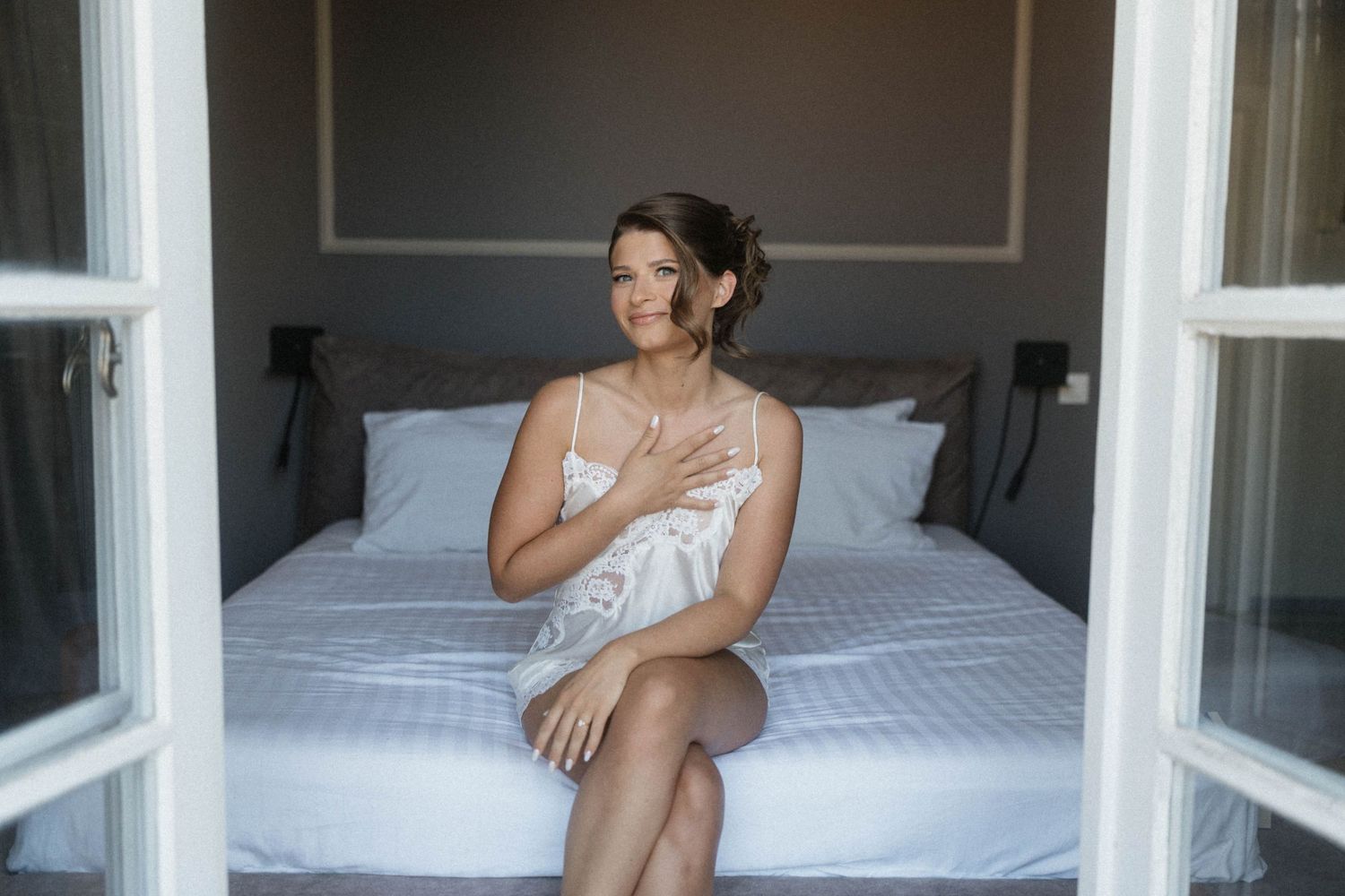 Elegant bride in white lace lingerie sitting on a bed, gazing thoughtfully in a softly lit bedroom.