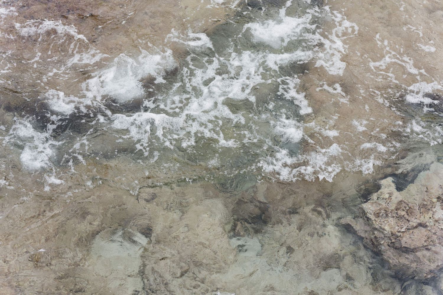 Aerial view of clear shallow water with white foam and ripples washing over submerged rocks and sandy seabed along a rocky shoreline.
