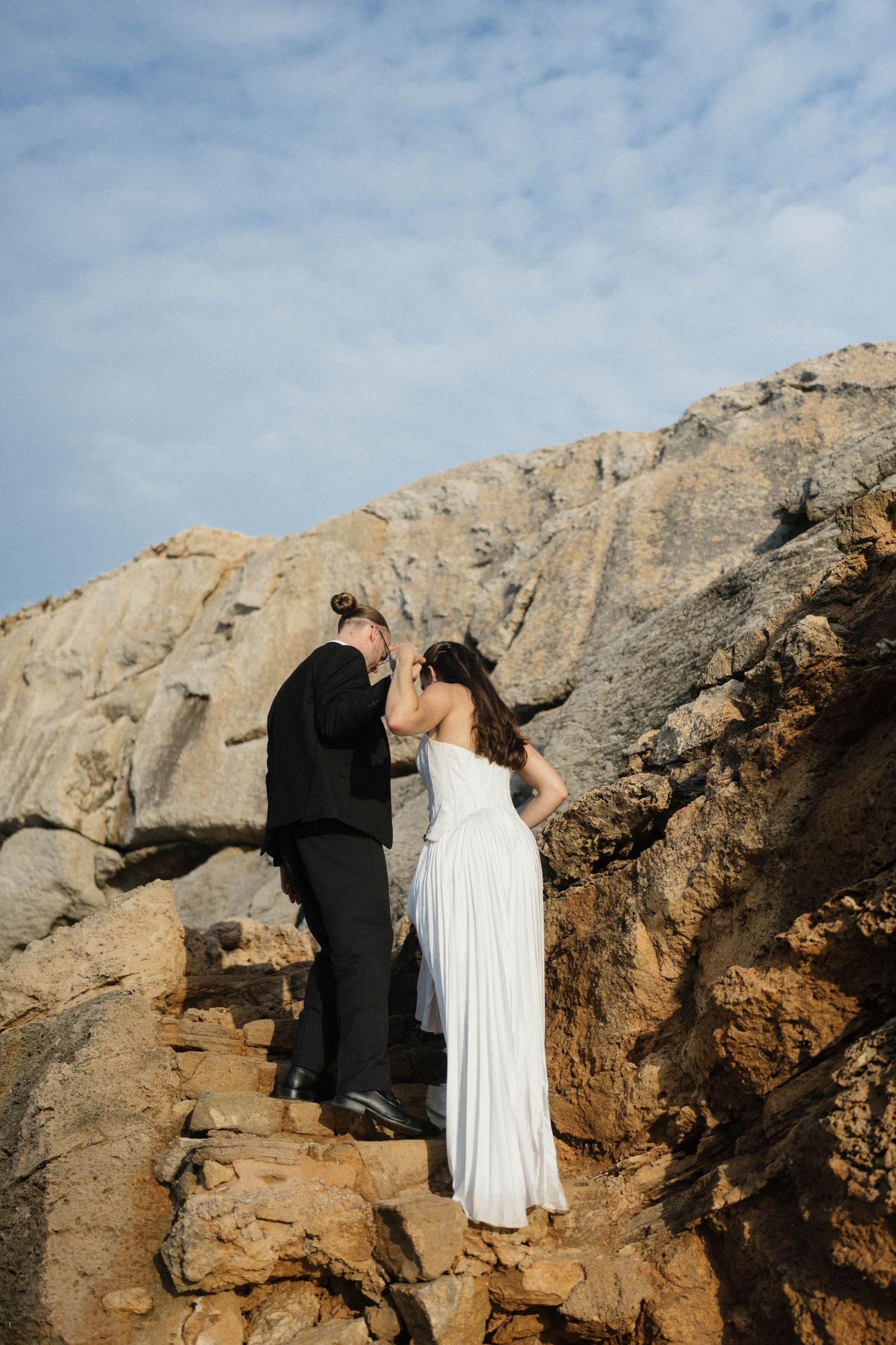Bride in a flowing white gown and groom in a black suit climb a rocky coastal staircase beneath a blue sky.