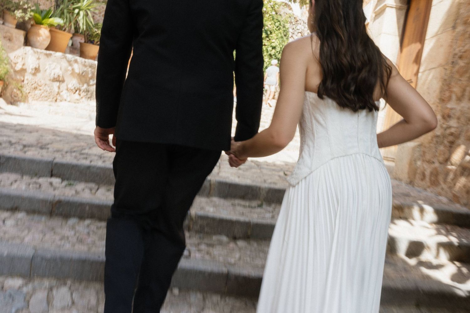 Bride in a white dress and groom in a dark suit hold hands as they walk up stone steps on a sunny day.
