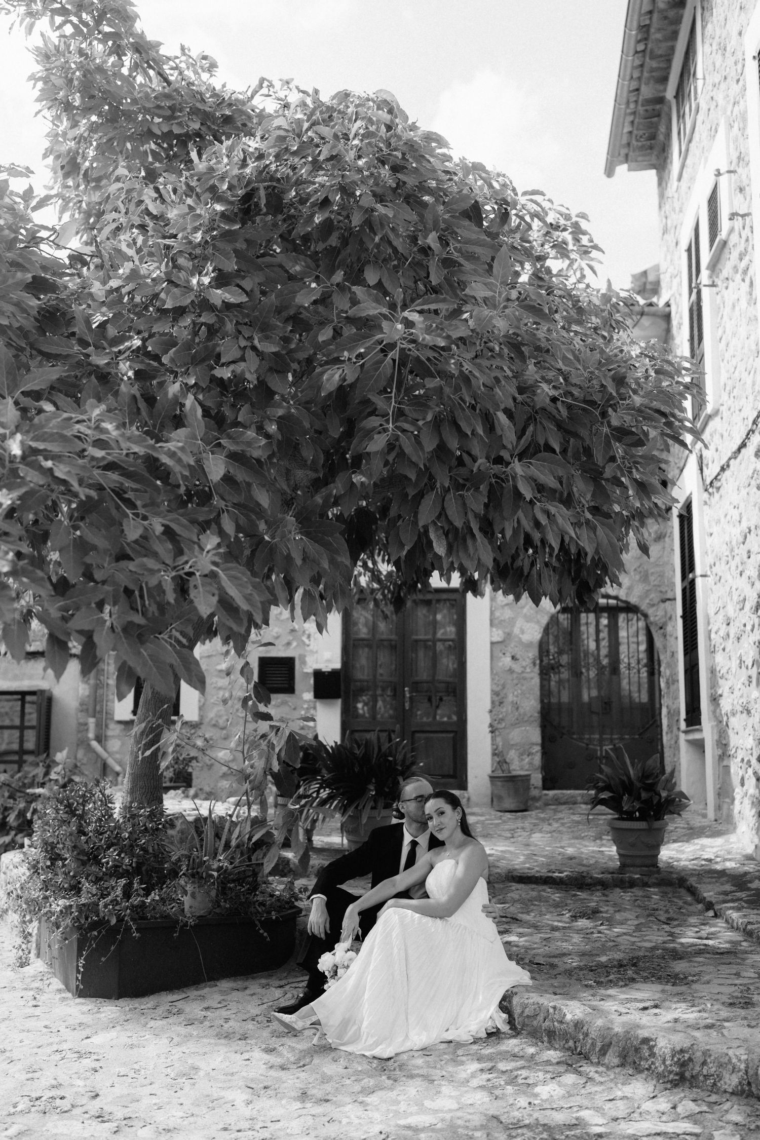 Black and white wedding photo of a bride and groom sitting together on a stone step beneath a leafy tree in a rustic cobblestone village.