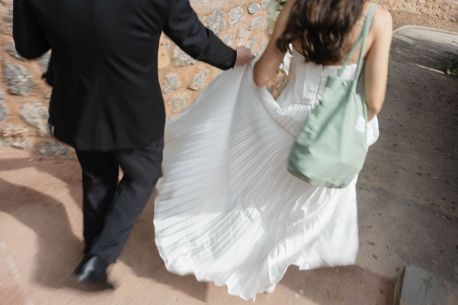 Bride in a flowing white pleated gown holds hands with a groom in a dark suit as they walk along a rustic stone path.