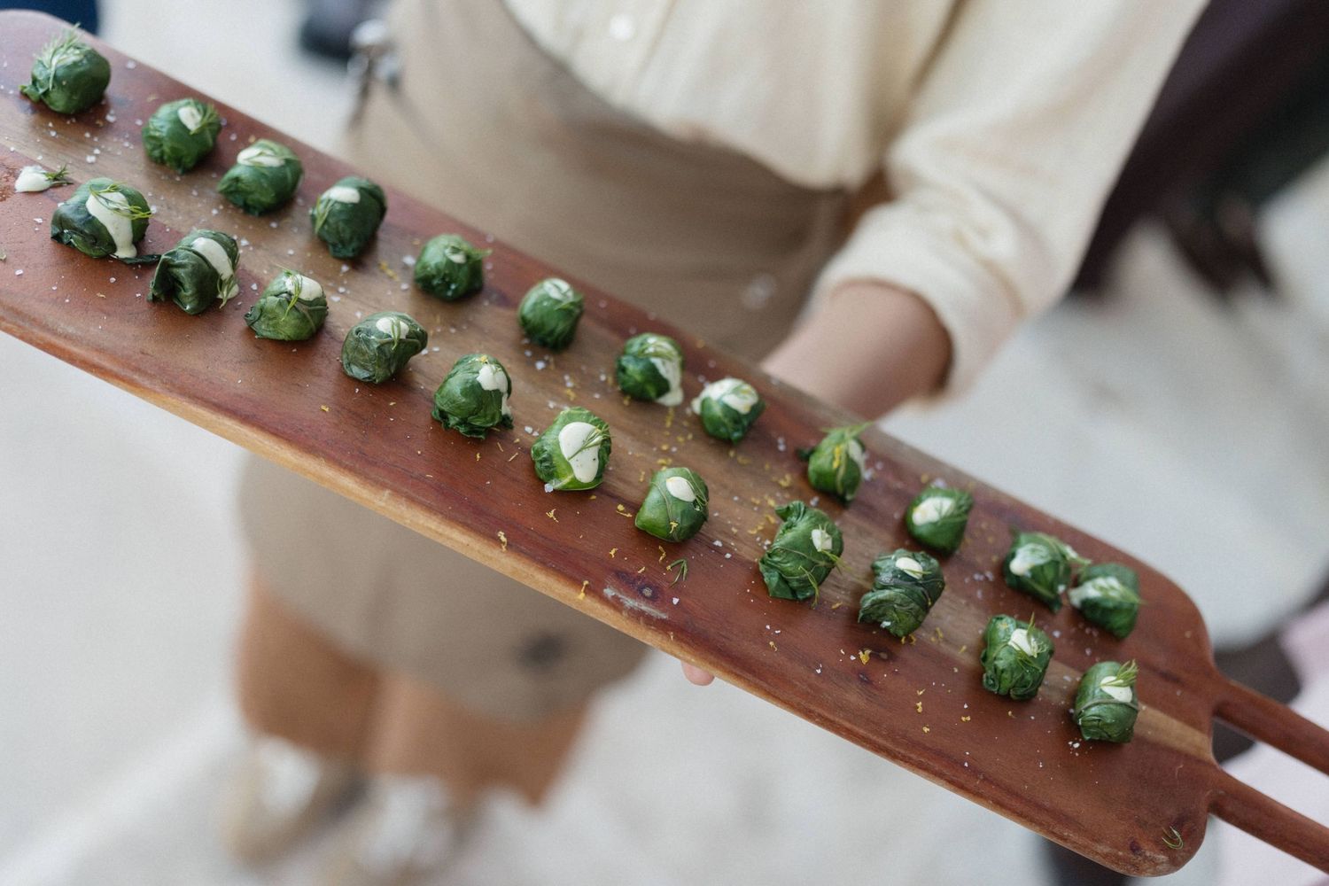 Person in a beige apron holds a long wooden paddle serving rows of seasoned, glossy stuffed grape leaves or Brussels sprouts appetizers.