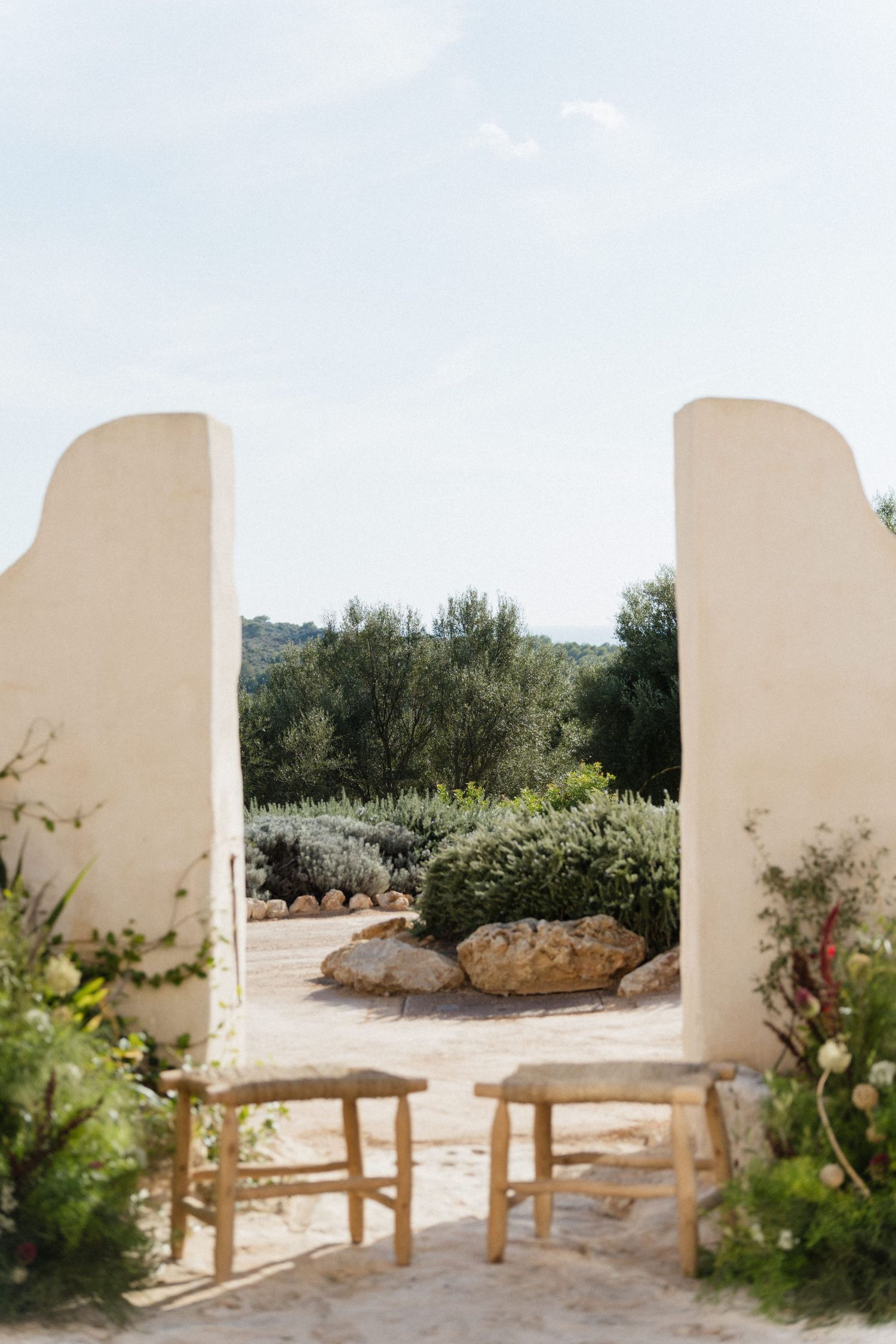 Two wooden stools and floral arrangements frame a whitewashed archway overlooking a Mediterranean garden path.