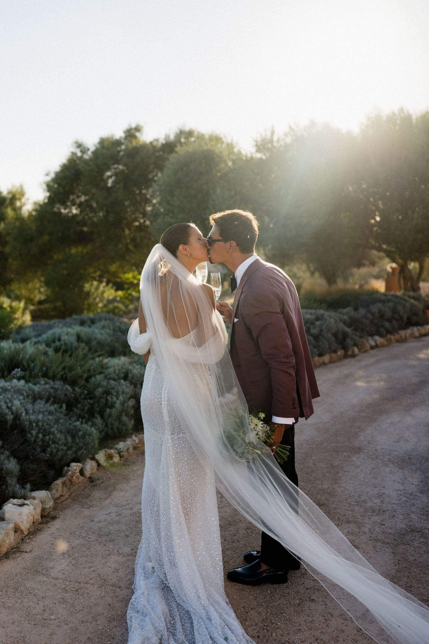 A bride in a sparkling gown with a flowing veil kisses her groom in a burgundy suit on a sunlit path lined with olive trees.