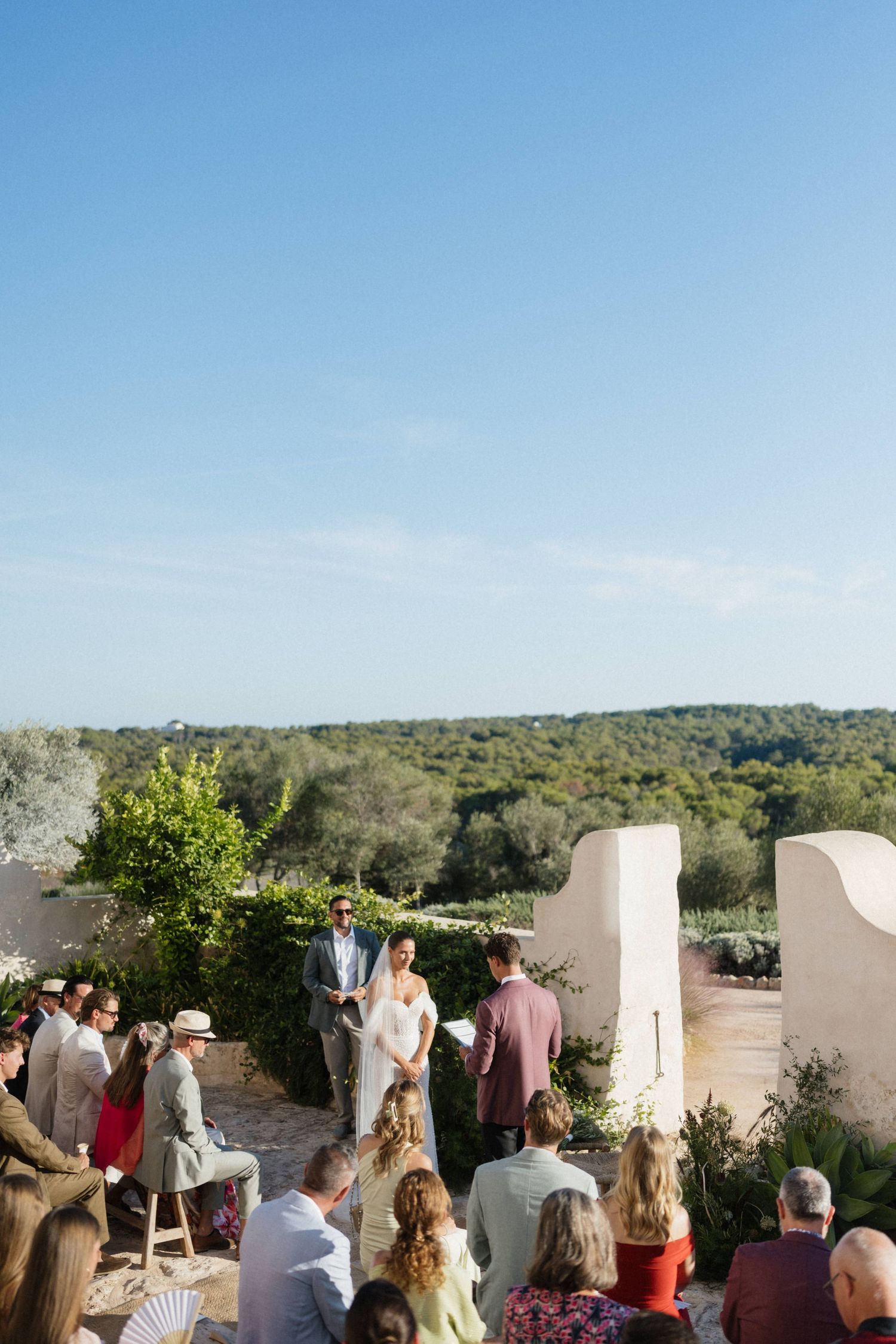 Wedding ceremony with the couple before an officiant, guests seated in the sun and green countryside stretching behind.