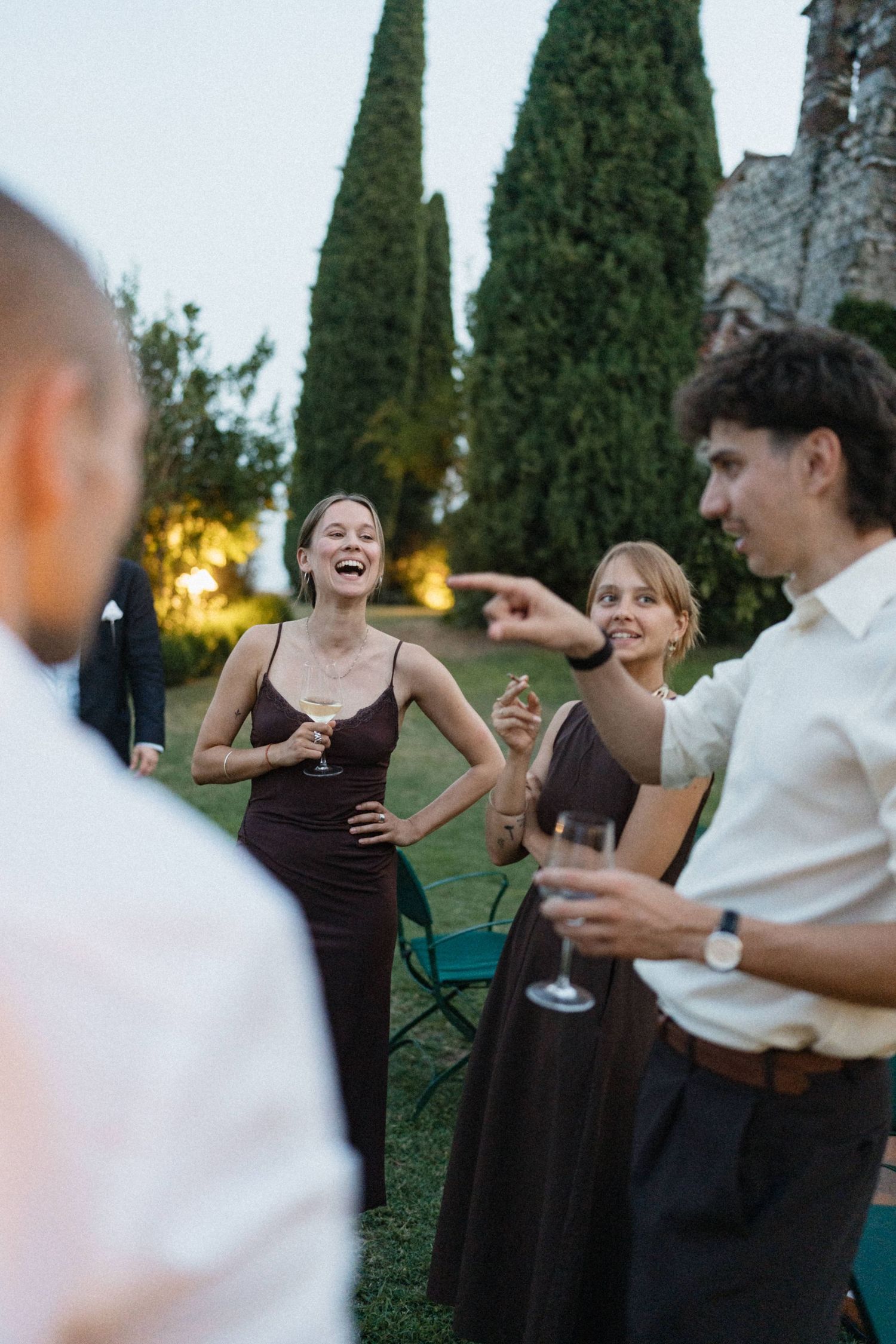 Elegantly dressed guests laughing and socializing at an outdoor evening garden party with cypress trees in the background.