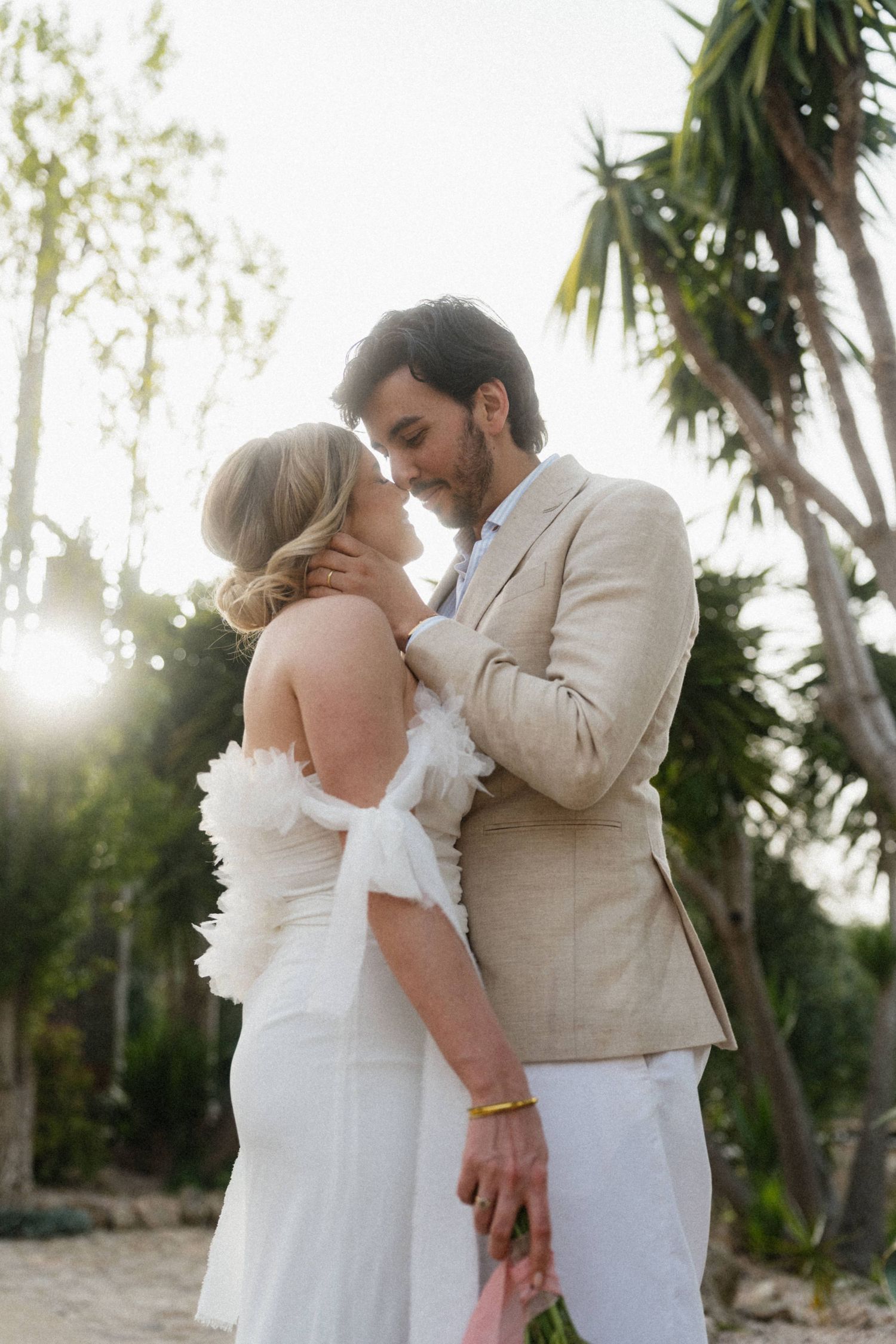 Bride and groom sharing a romantic moment surrounded by palm trees in golden backlit sunlight.