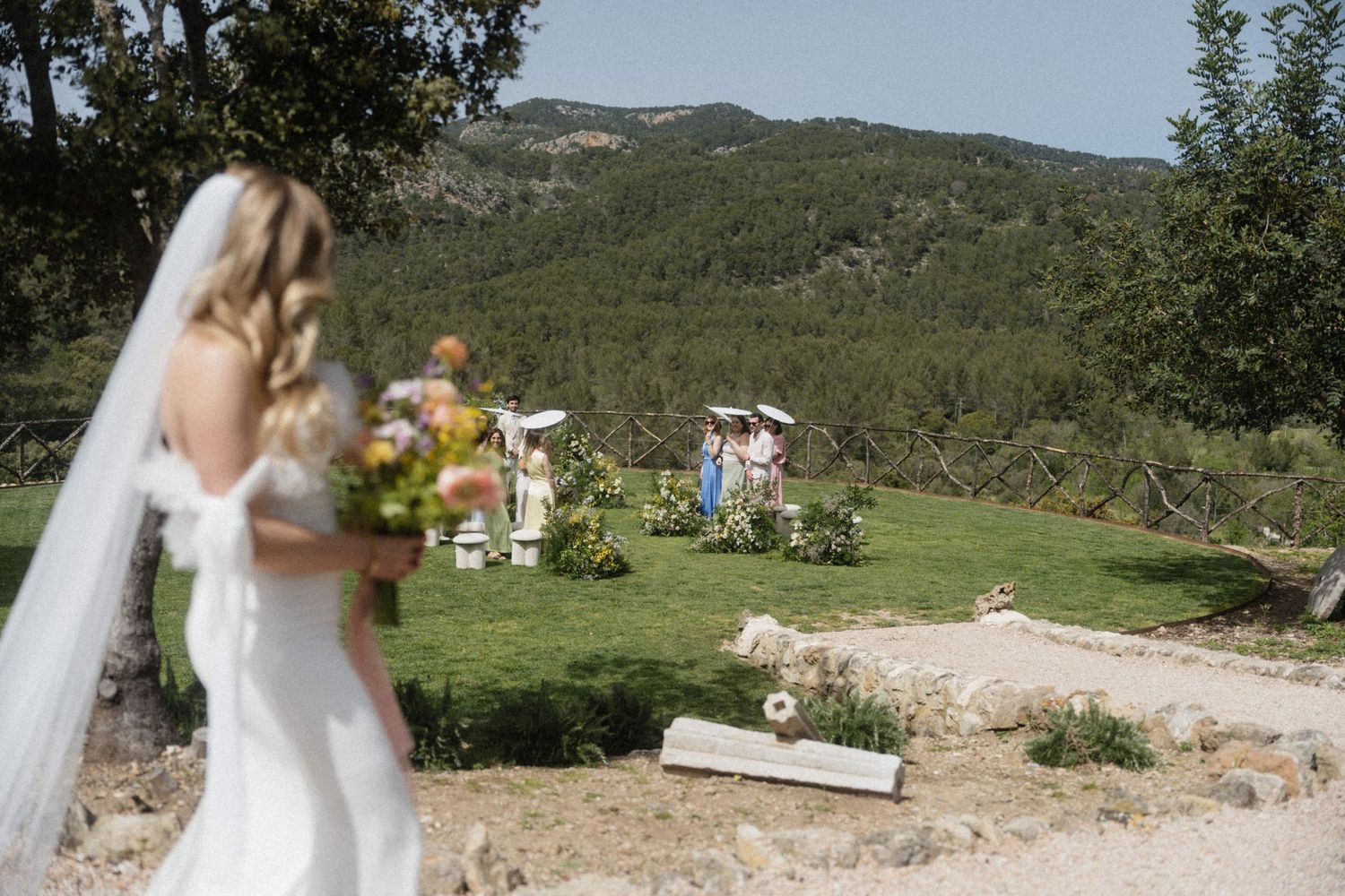 Blonde bride in white gown with veil holding bouquet overlooking outdoor mountain wedding ceremony.