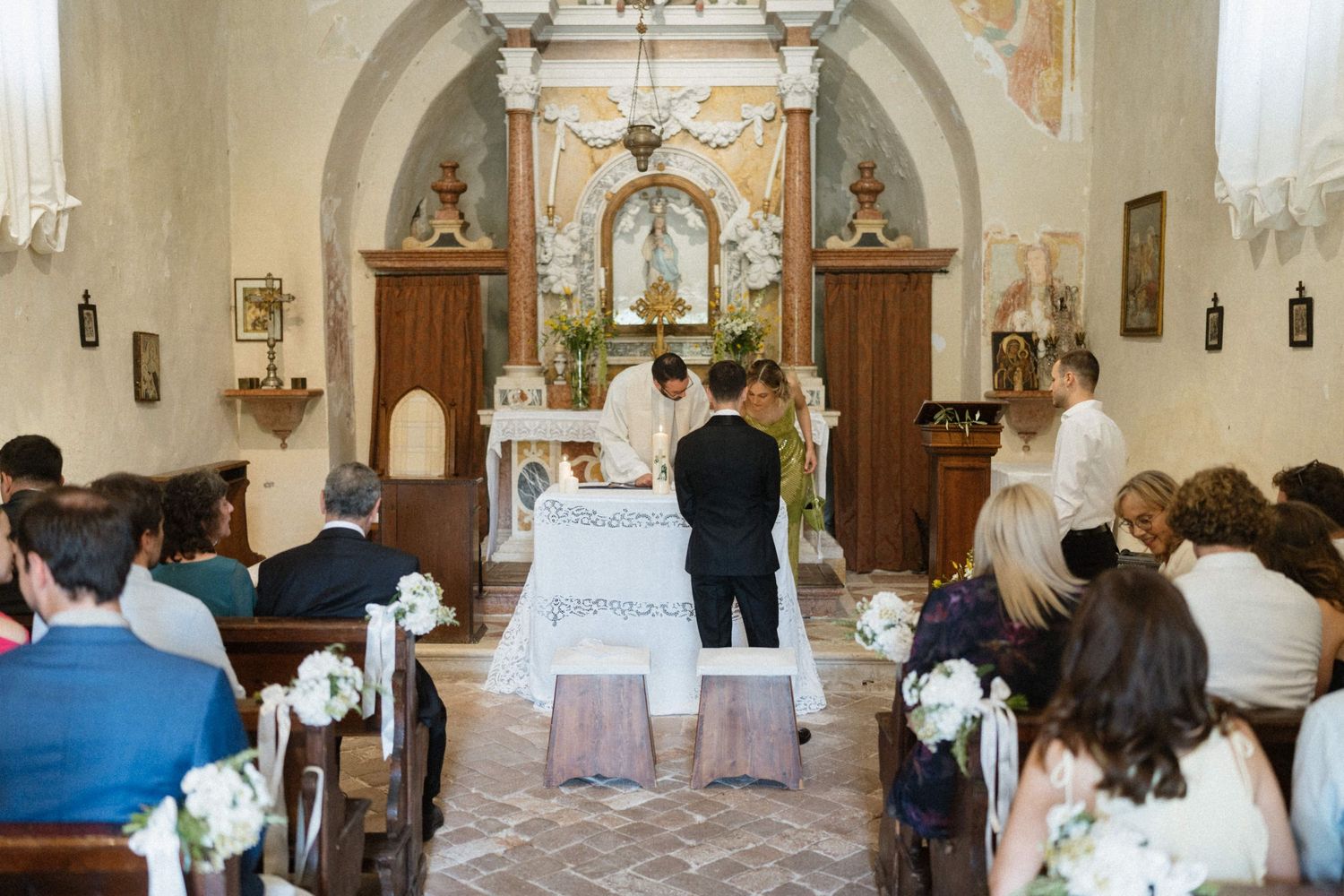 Couple exchanging vows at a rustic Italian church altar, surrounded by guests seated in wooden pews during a wedding ceremony.