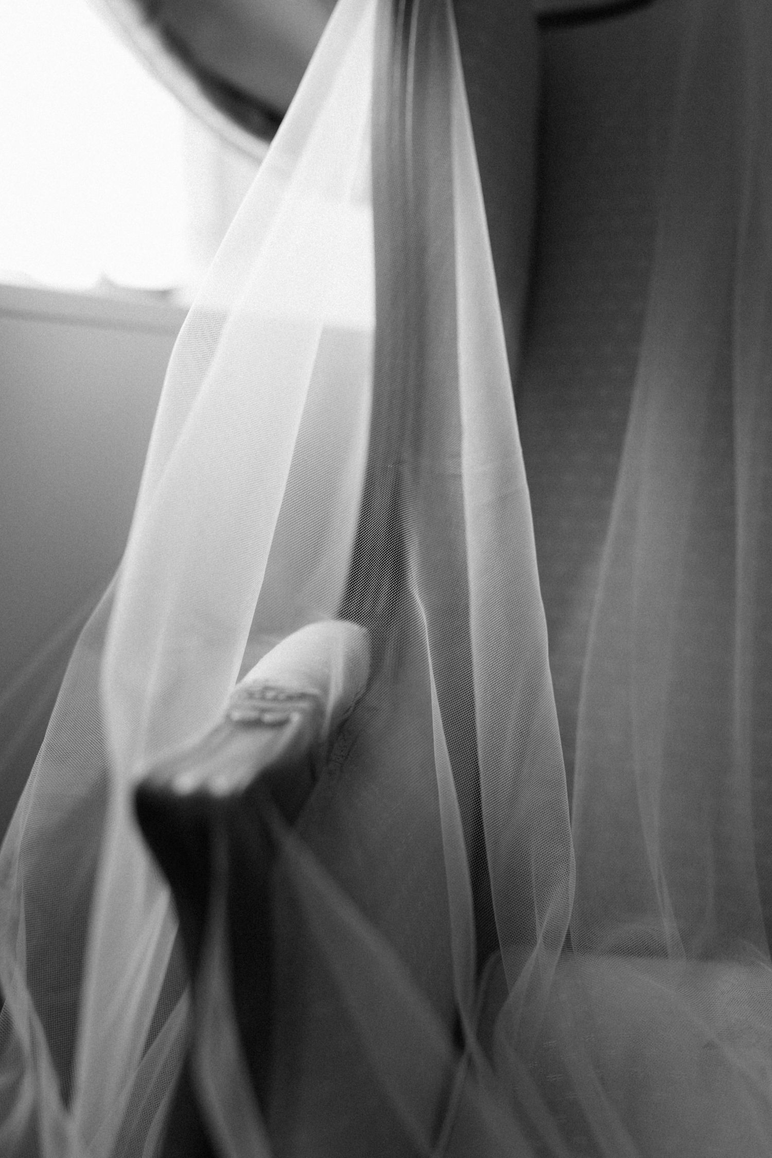 Black and white photo of a sneaker peeking beneath a flowing bridal veil near a sunlit window.