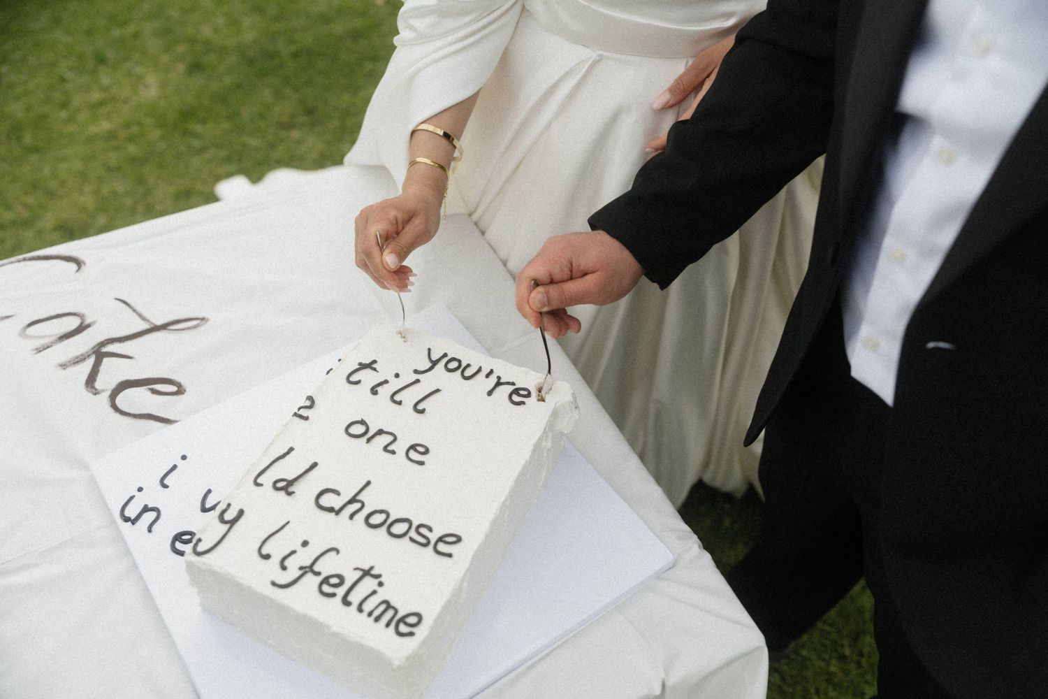 Wedding couple cutting white cake inscribed with romantic vow lyrics on outdoor grass setting.