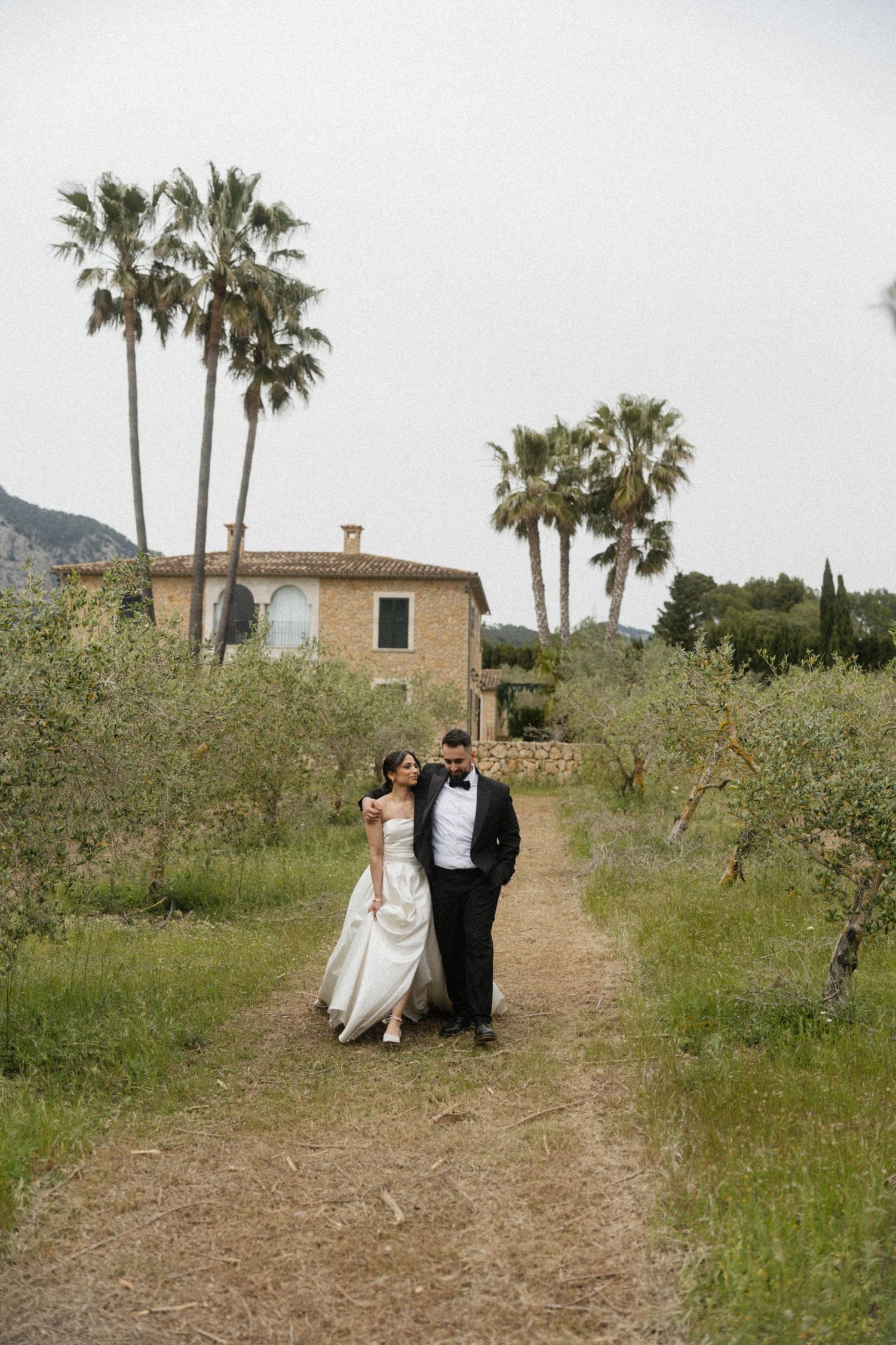 Bride and groom walking along dirt path with palm trees and rustic stone villa behind them.
