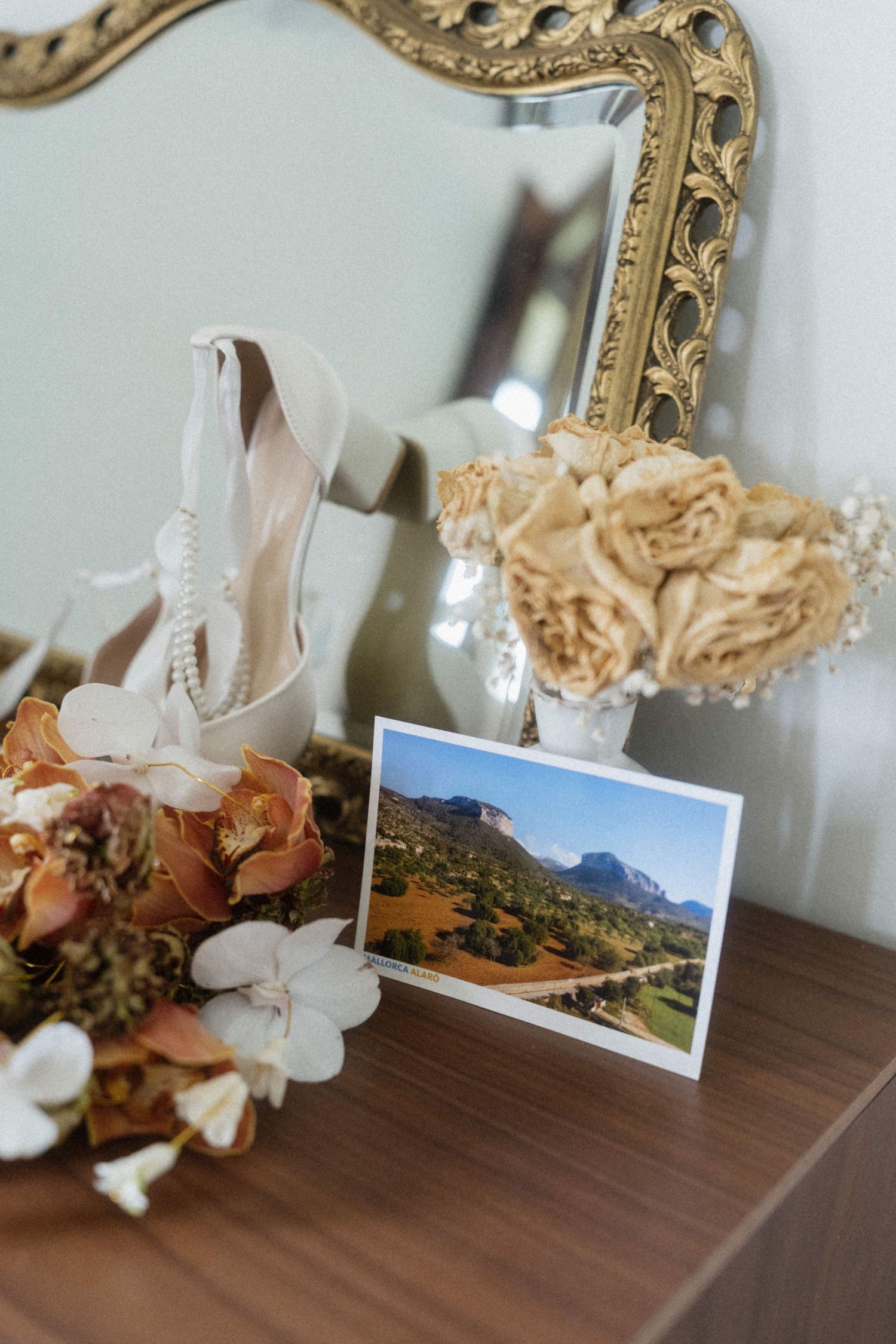 Wedding details flat lay with bridal heels, floral bouquet, and postcard on wooden dresser with ornate mirror.