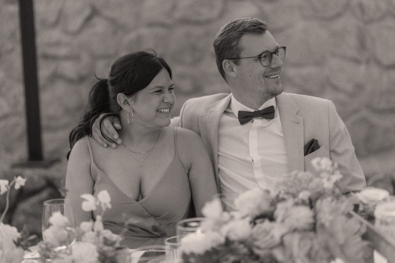 Black and white photo of a smiling couple at a wedding reception table, groom in bow tie and suit, bride in elegant dress.