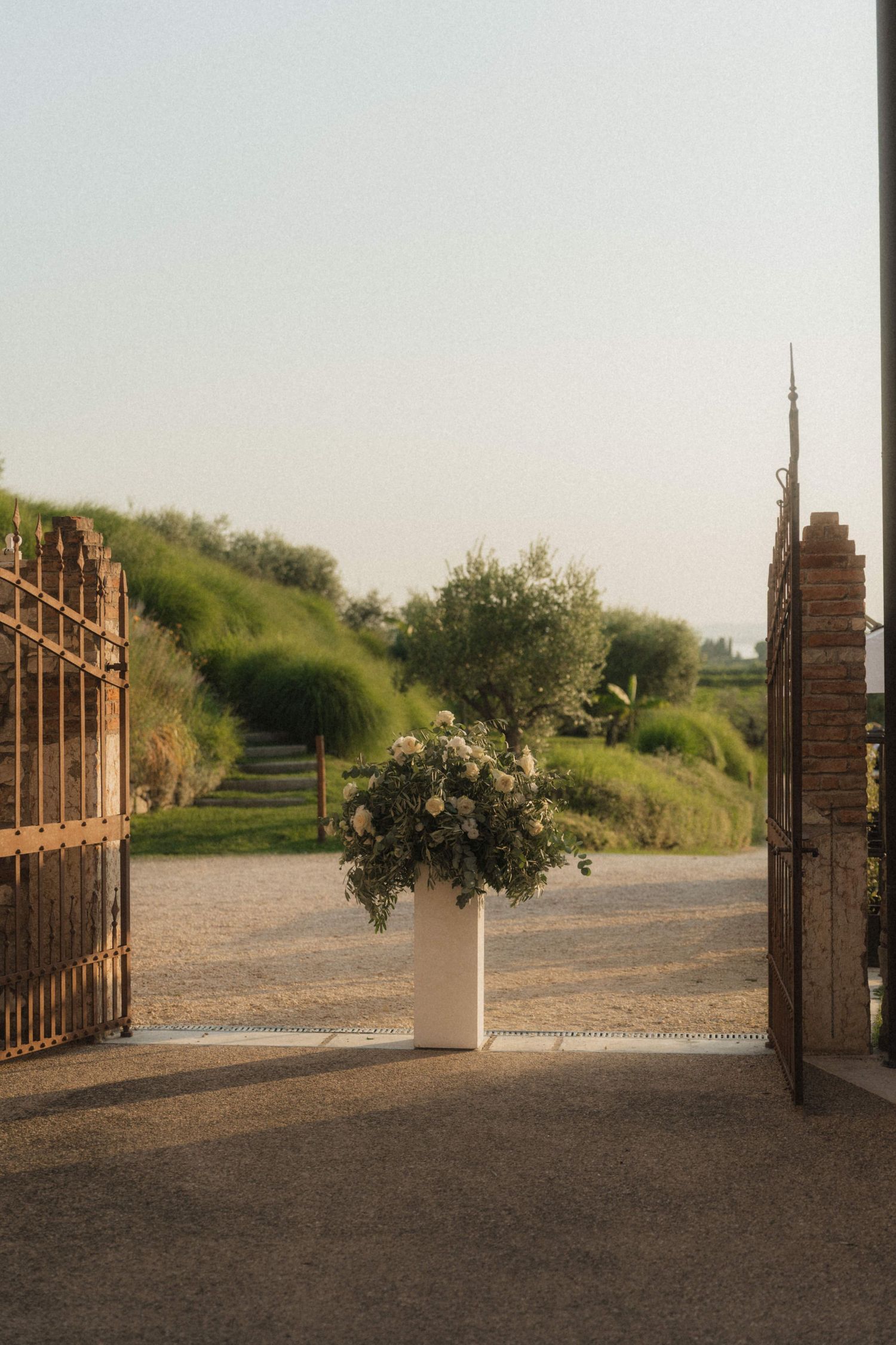 White floral arrangement in planter framed by rustic iron gates at wedding venue entrance.