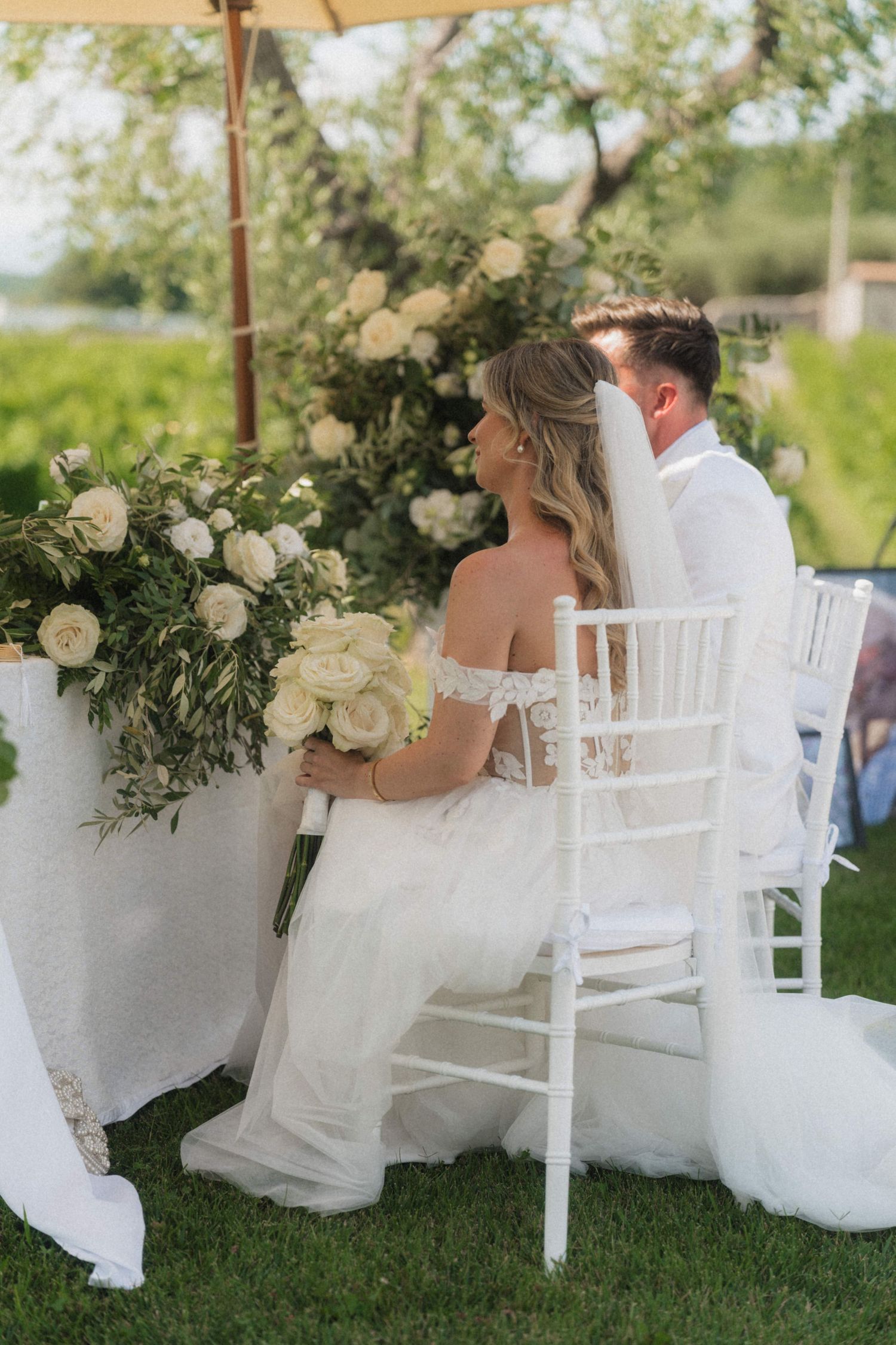 Bride and groom sitting together at outdoor wedding ceremony with white floral arrangements and lush greenery.
