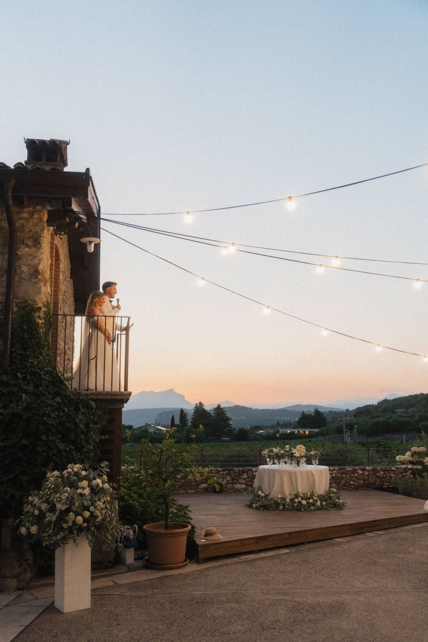 Bride standing on stone villa balcony at dusk with string lights and mountain views.