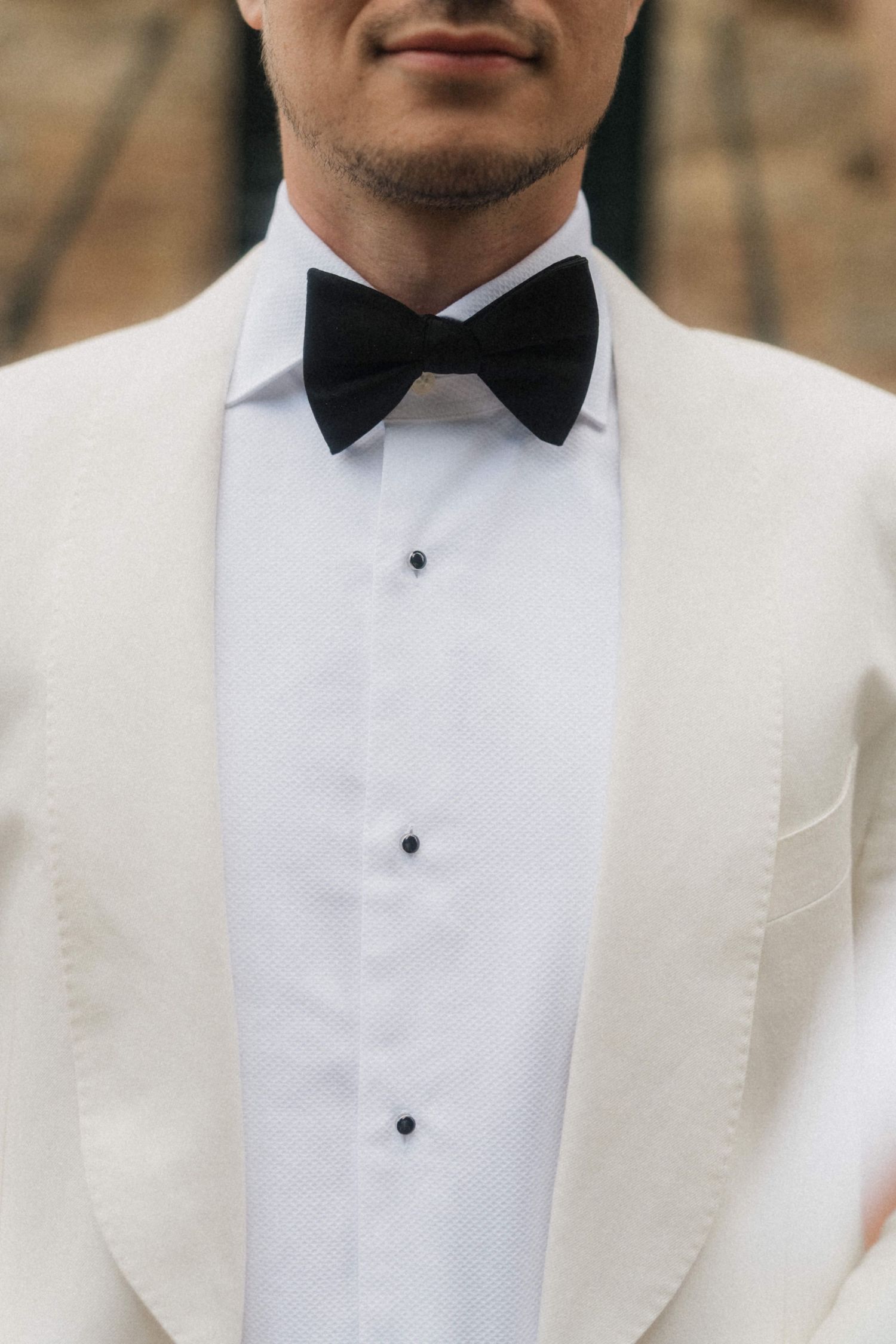 Groom in white tuxedo jacket with black bow tie and dress shirt at wedding ceremony.