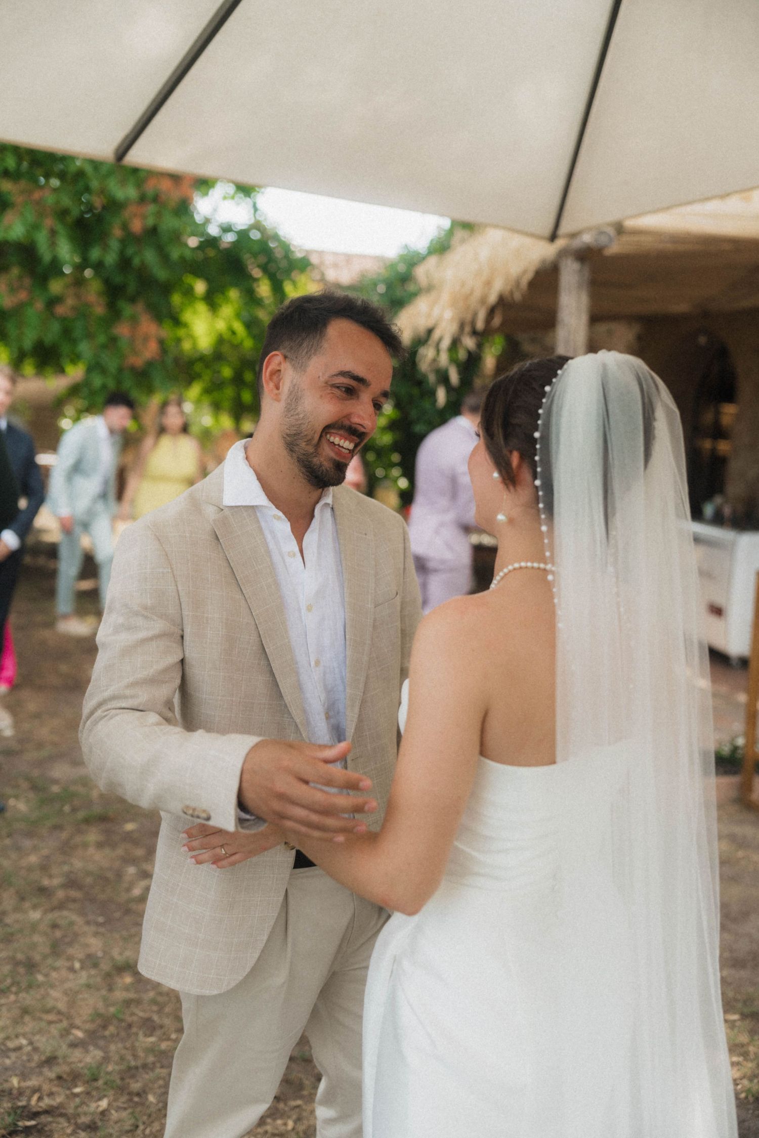 Smiling groom in beige suit greets bride in white dress and veil at outdoor wedding reception.