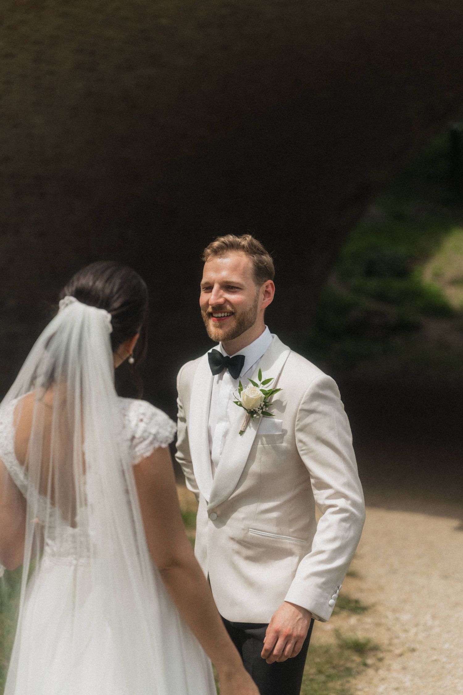 Smiling groom in white tuxedo with boutonniere sharing first look moment with bride in veil outdoors.