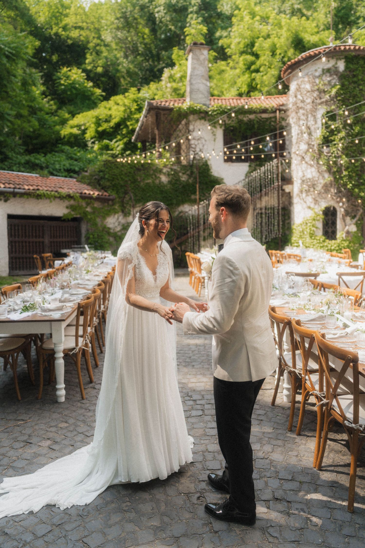 Bride in flowing white gown and groom in ivory tuxedo holding hands at an elegantly set outdoor wedding reception.