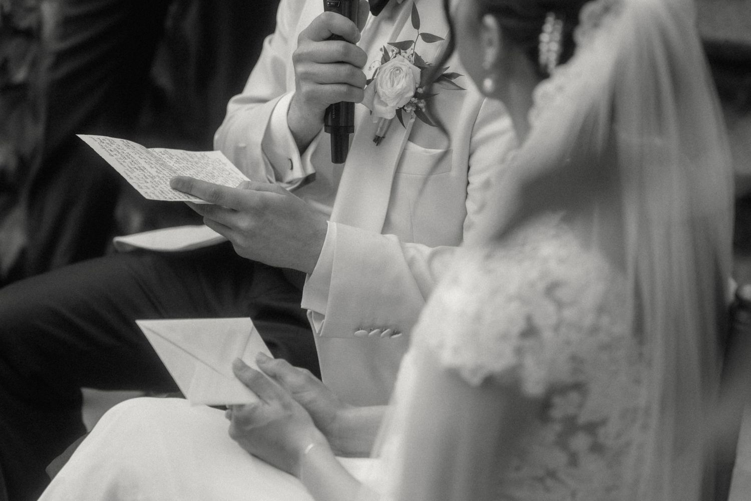 Black and white close-up of groom reading wedding vows while bride in lace dress holds paper.