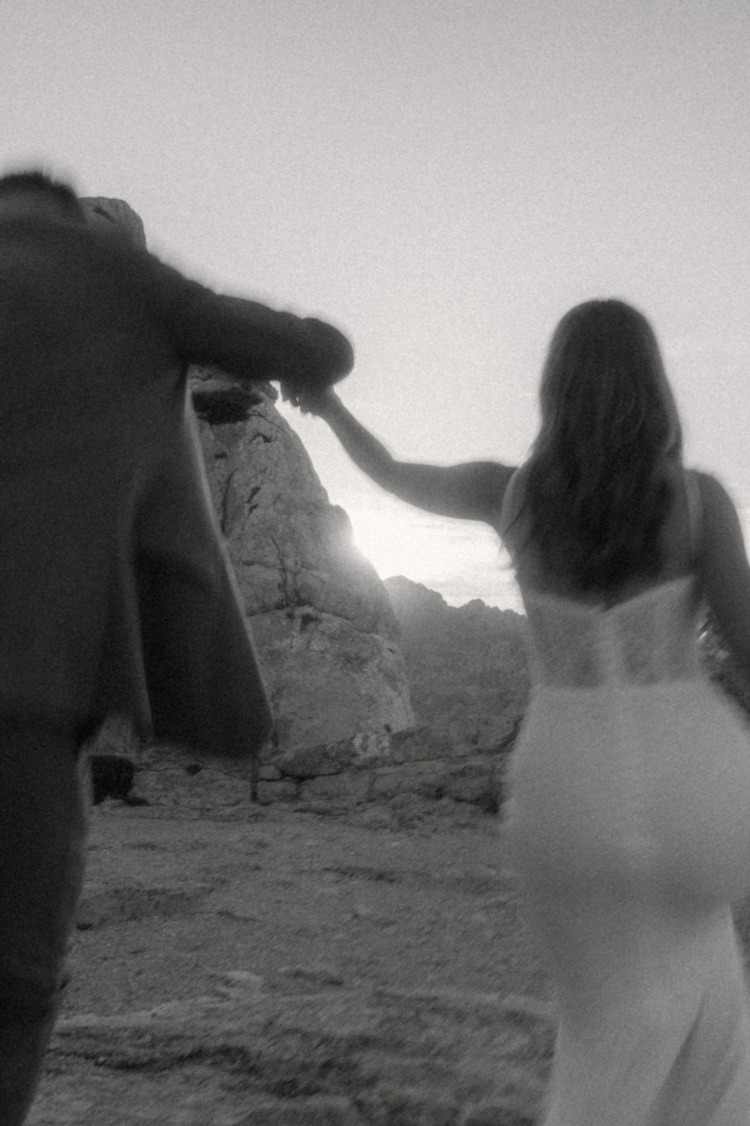 Artistic blurred black and white shot of bride and groom walking hand in hand toward rocky cliffs at sunset.