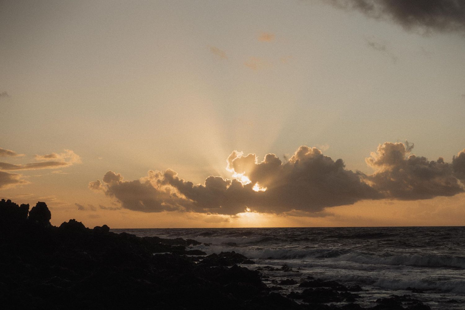 Golden sunset with dramatic sun rays bursting through clouds over rocky ocean coastline.