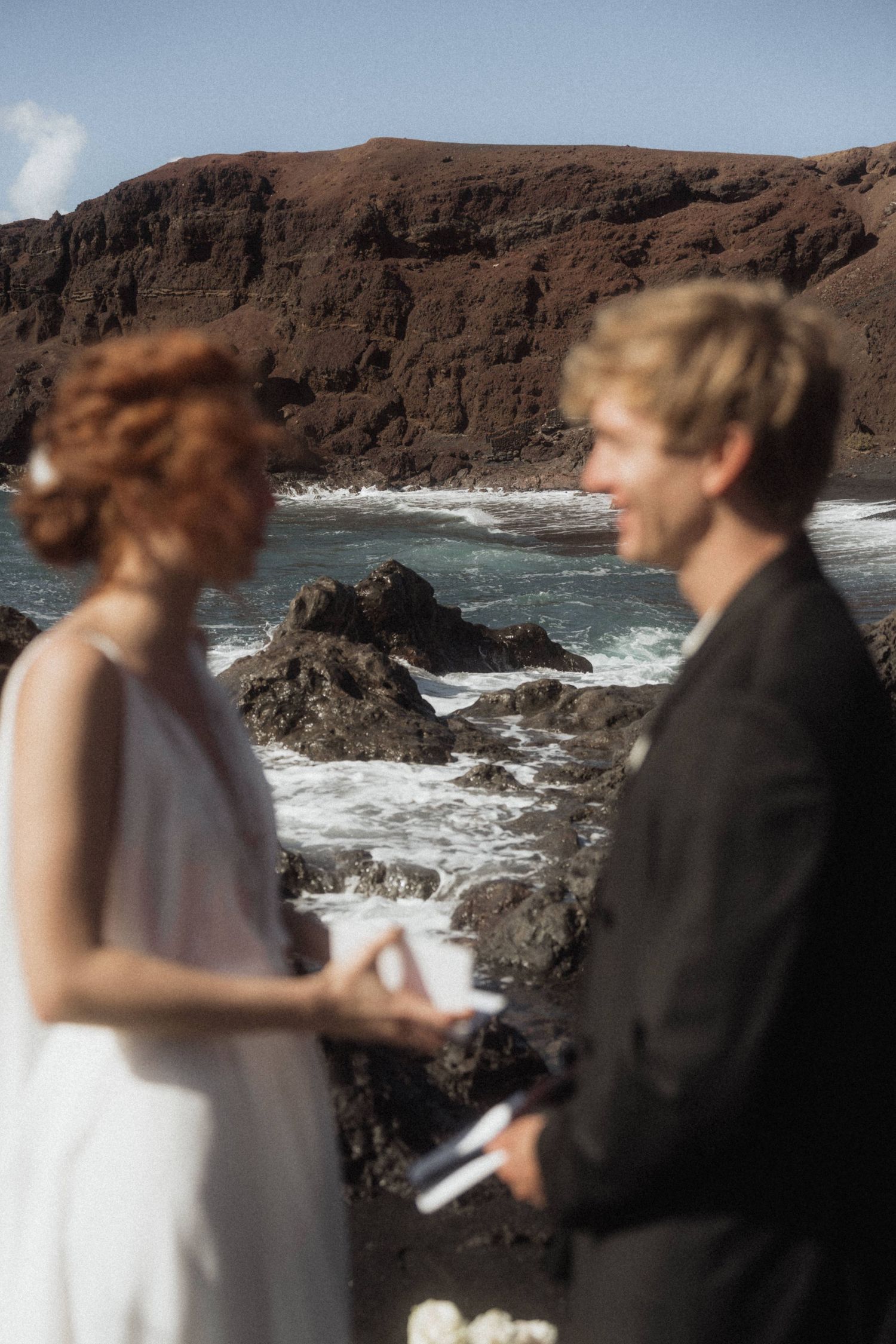 Couple exchanging vows at dramatic volcanic black rock coastline with crashing ocean waves behind them.