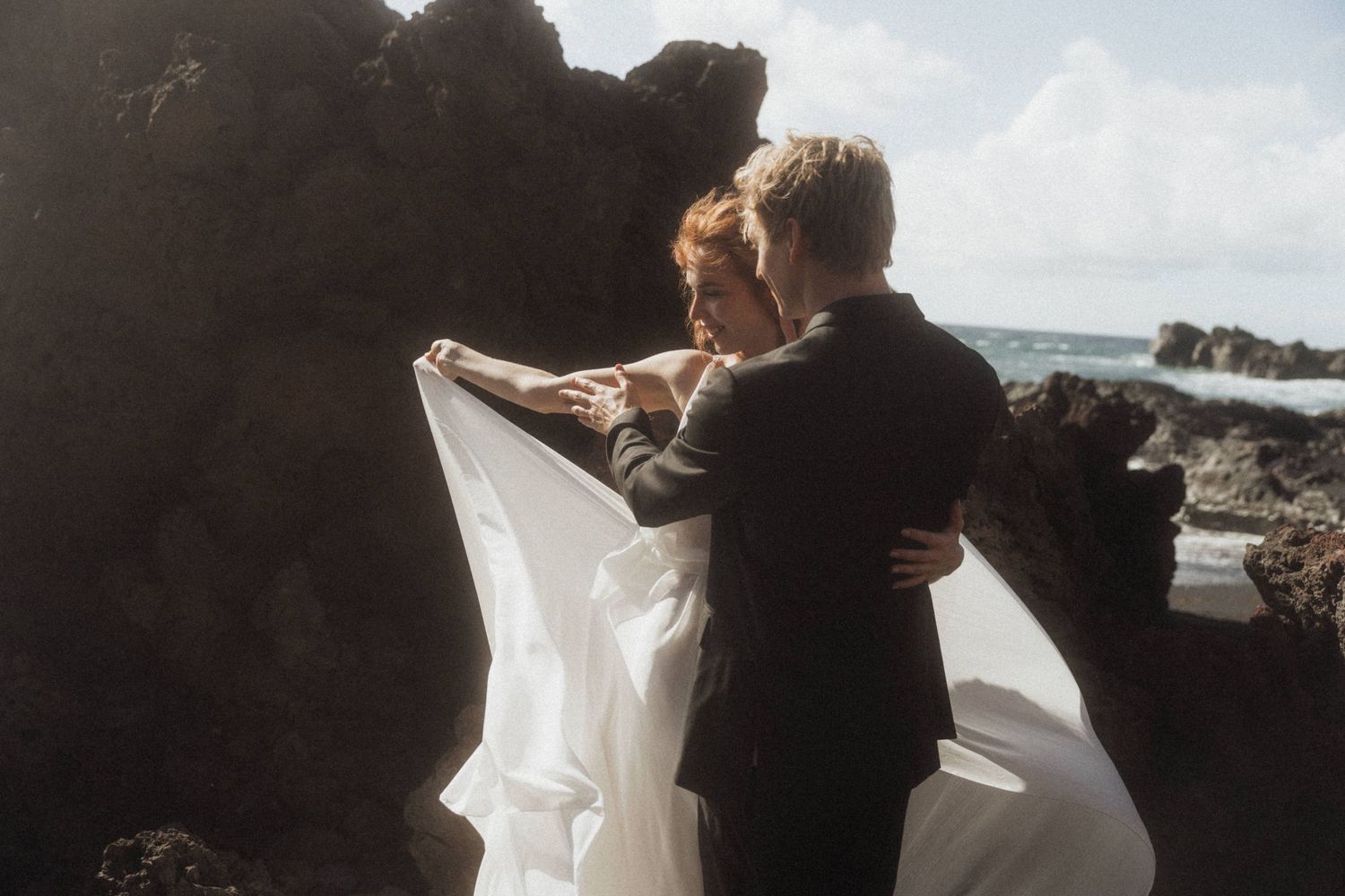 Couple embraces among dark volcanic rocks as bride's white veil flows in ocean breeze.