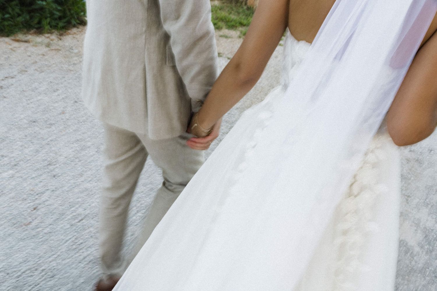 Close-up of bride in white lace gown and groom in beige suit holding hands outdoors.