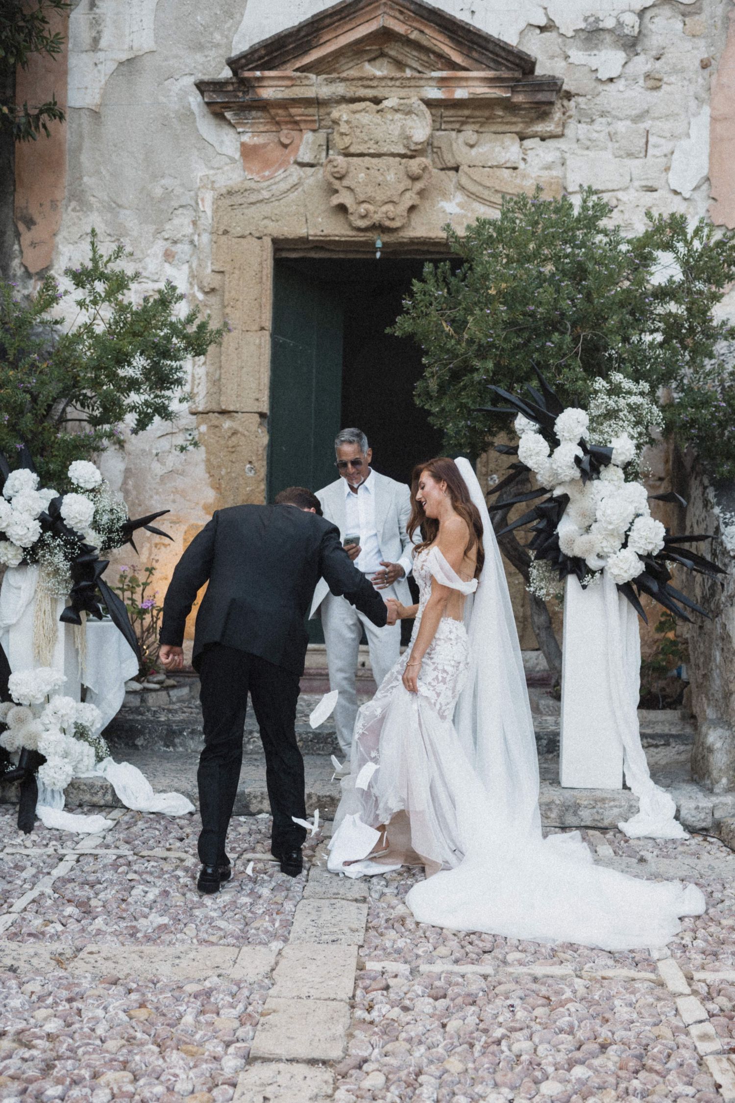 Groom adjusts bride's dress at rustic outdoor wedding ceremony with floral decor.