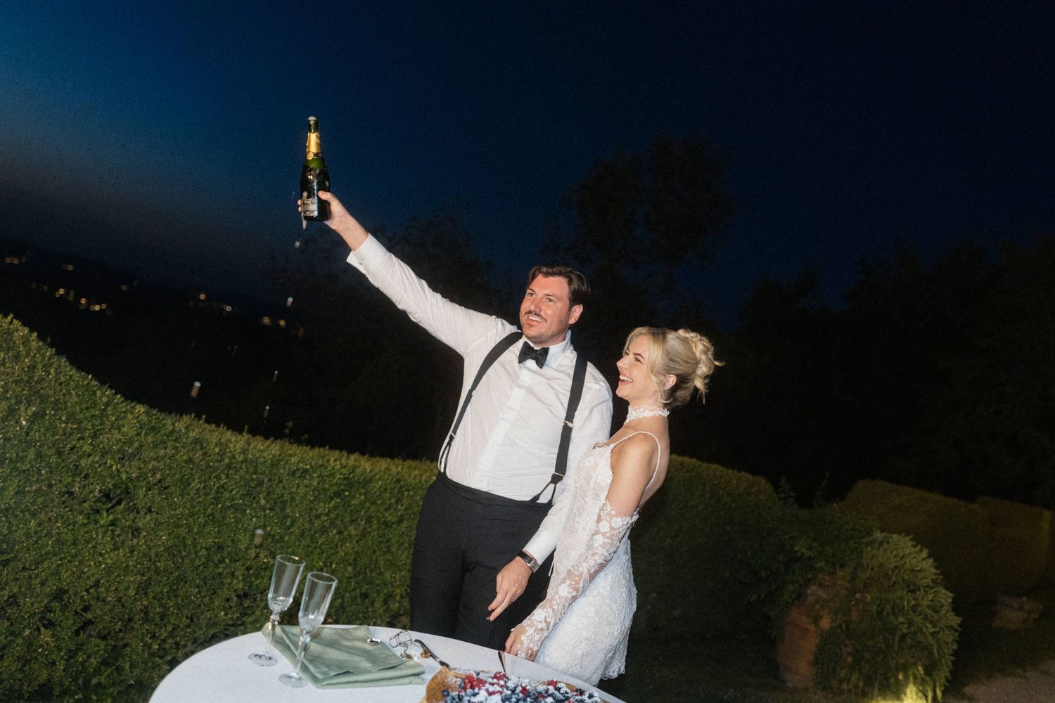 Groom raising champagne bottle in celebration with bride at outdoor wedding reception at night.
