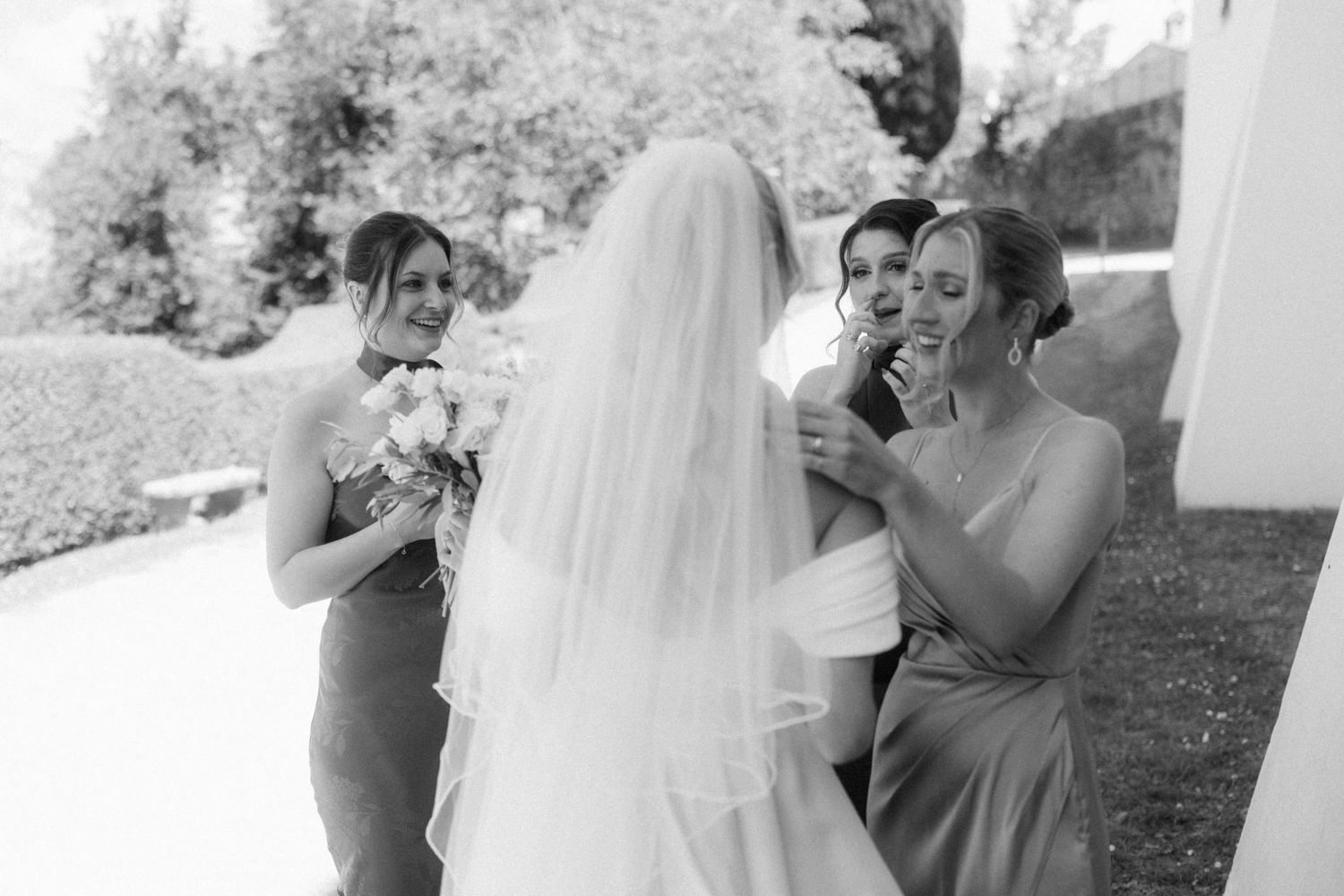 Bride in white veil shares emotional moment with smiling bridesmaids holding bouquets outdoors.