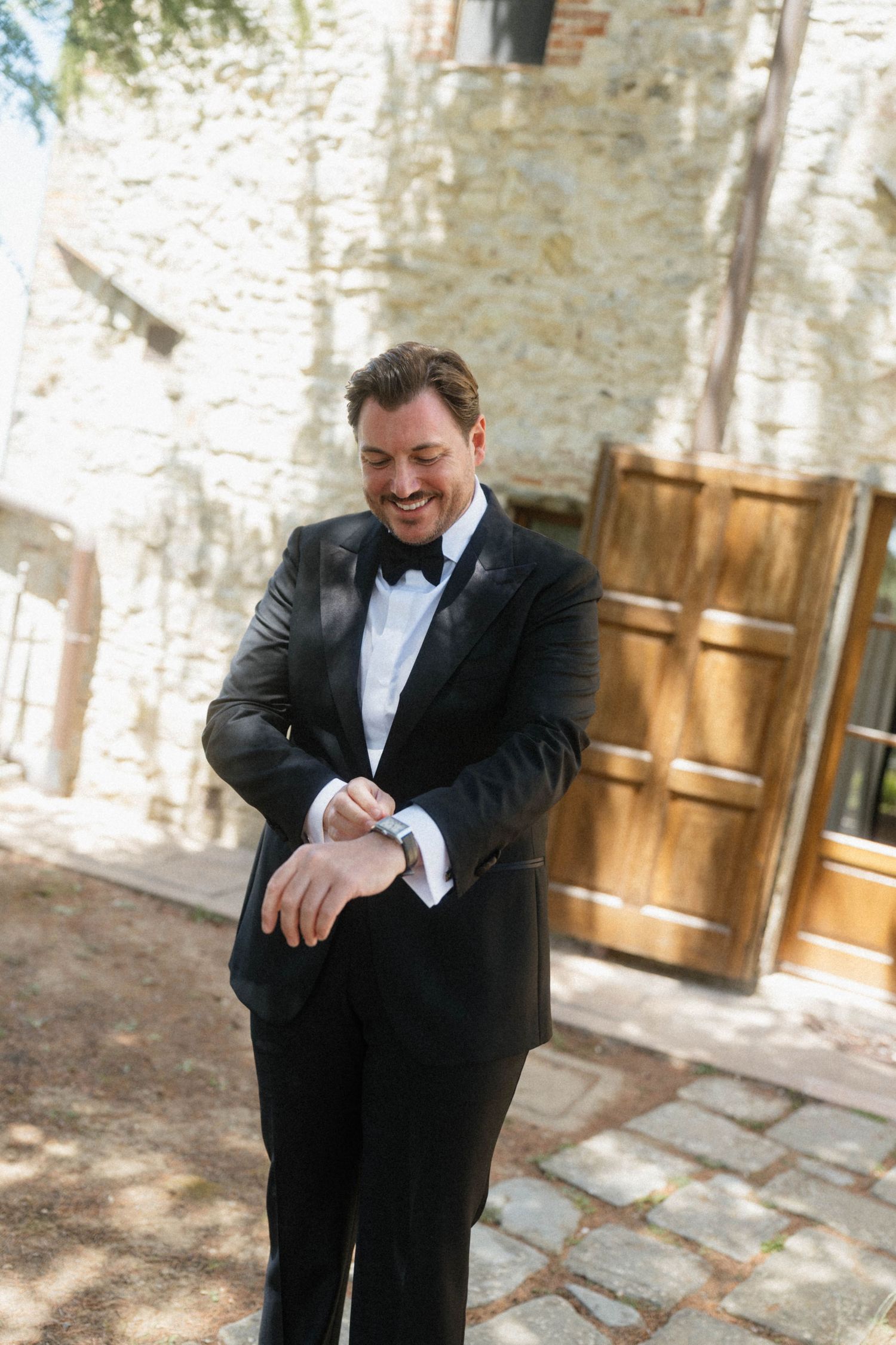 Smiling groom in black tuxedo checking his watch outside a rustic stone wedding venue.
